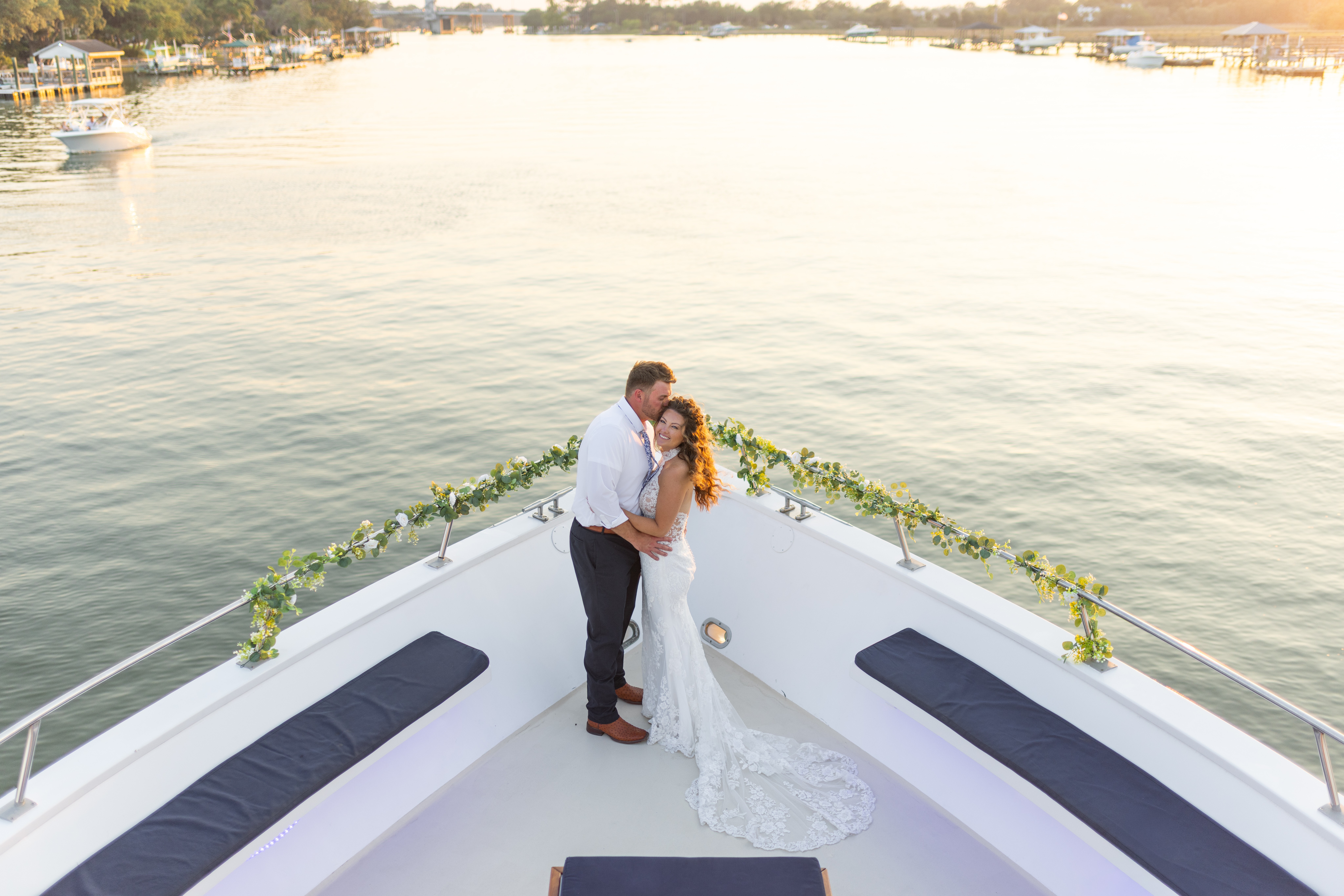A couple in wedding attire embraces on a boat with water and shoreline in the background.