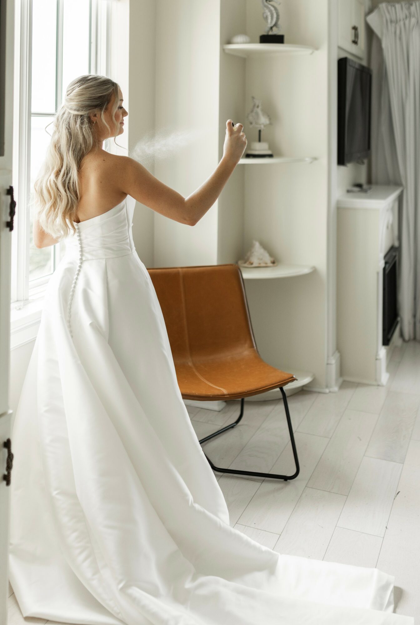 Woman in wedding dress reaching for an item on a white shelf, with a brown chair nearby, in a bright room.