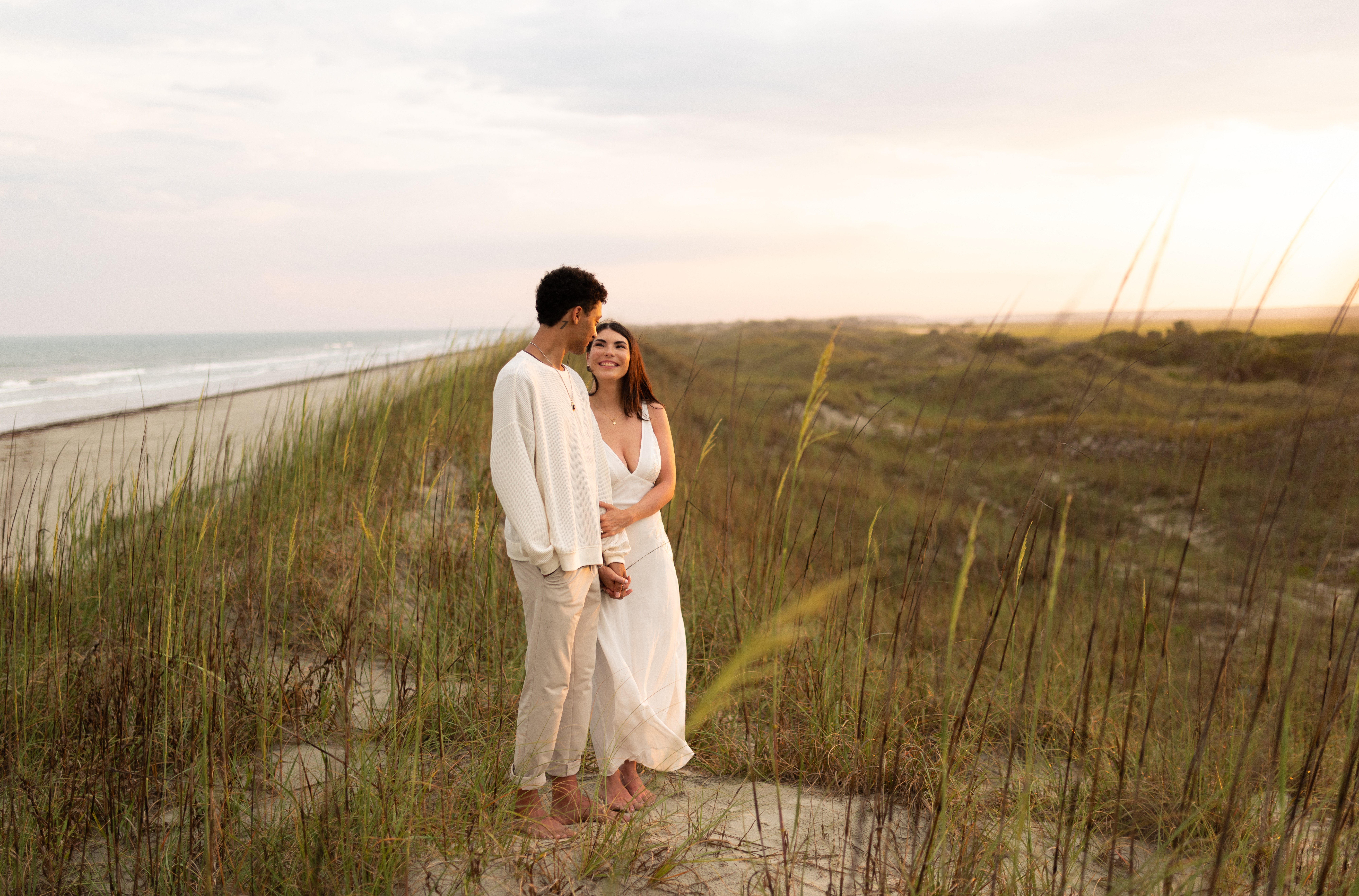 A couple stands on a beach path with grass, facing each other, during sunset, smiling and holding hands.