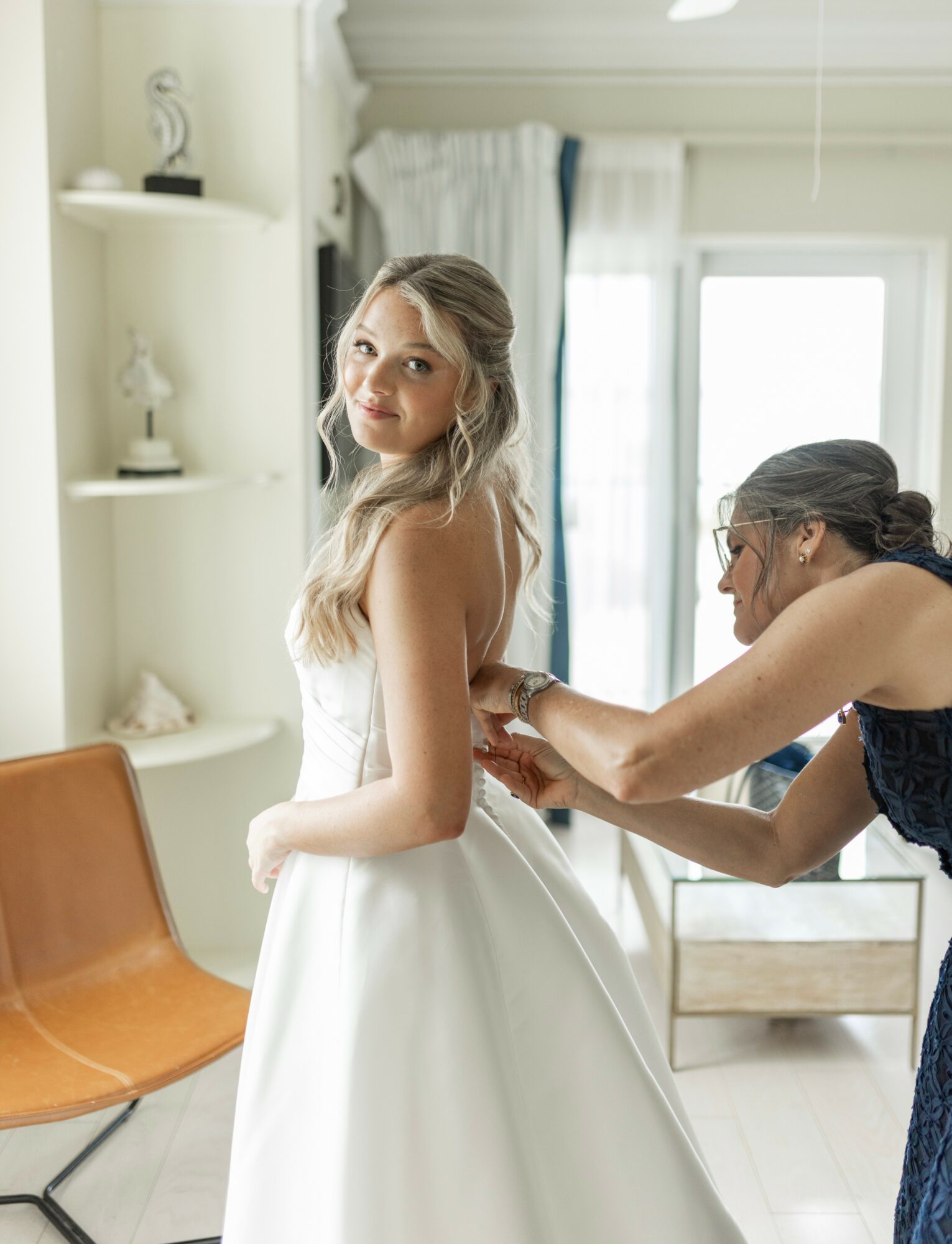Young woman in a white dress being assisted by another woman in a room with natural light, shelves, and a chair.