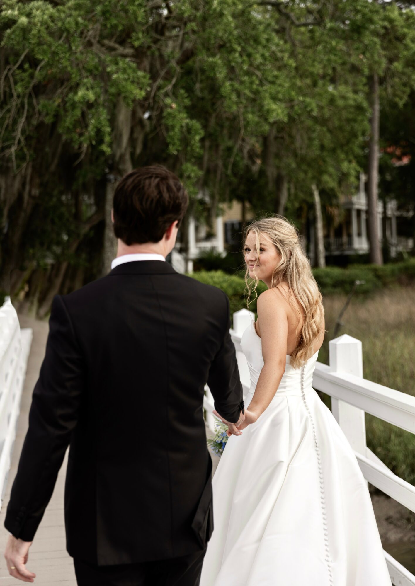 Bride and groom holding hands outdoors, bride in a white wedding dress, groom in a black suit, trees in background.