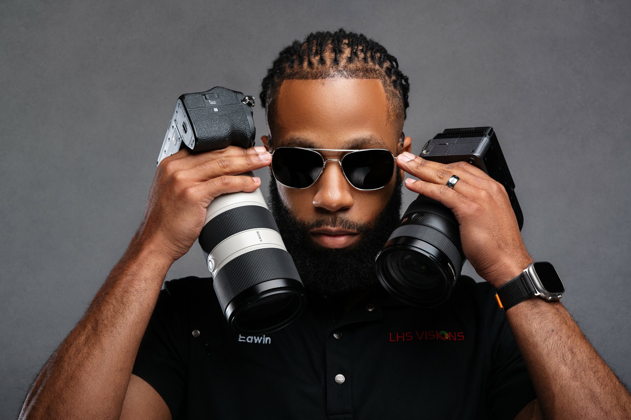 Man with sunglasses holding two cameras near his head, wearing a black shirt and watch, against a gray background.