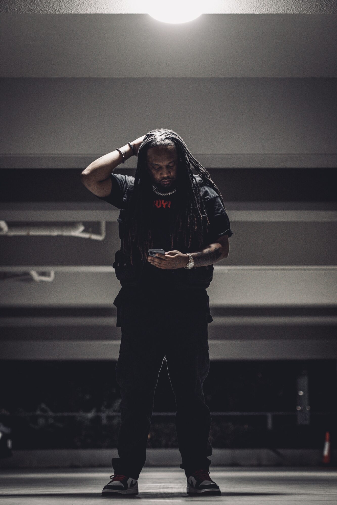 Person standing in a parking garage, looking at a phone, with one hand on their head, dim lighting.