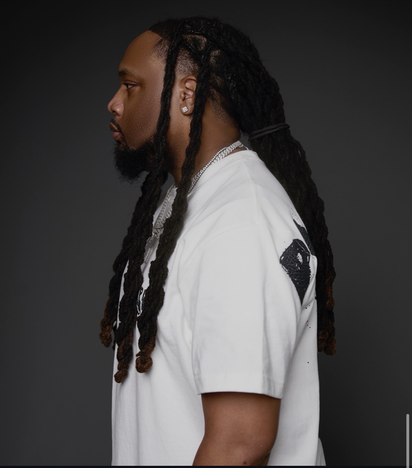 Side profile of a man with dreadlocks wearing a white T-shirt against a dark background.
