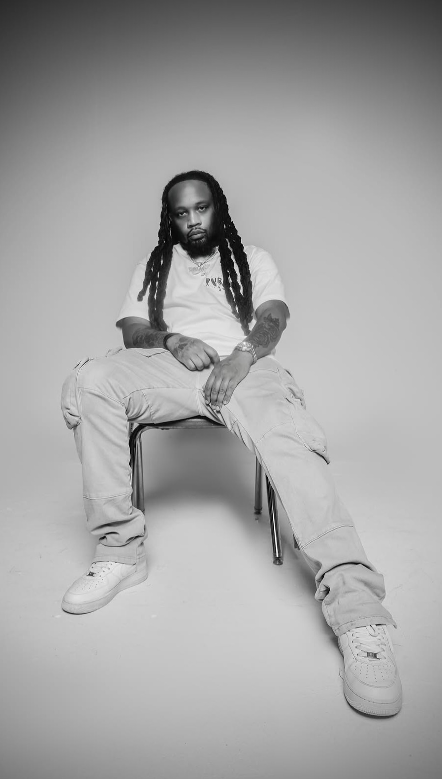 Person with long dreadlocks sitting on a chair, wearing a white shirt and light-colored pants, in a studio setting.