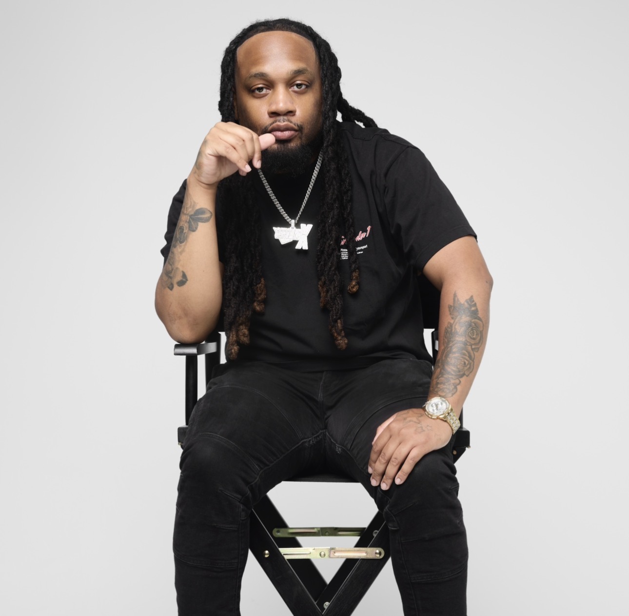 Man with long dreadlocks sitting on a chair, wearing black clothing and jewelry, against a plain background.