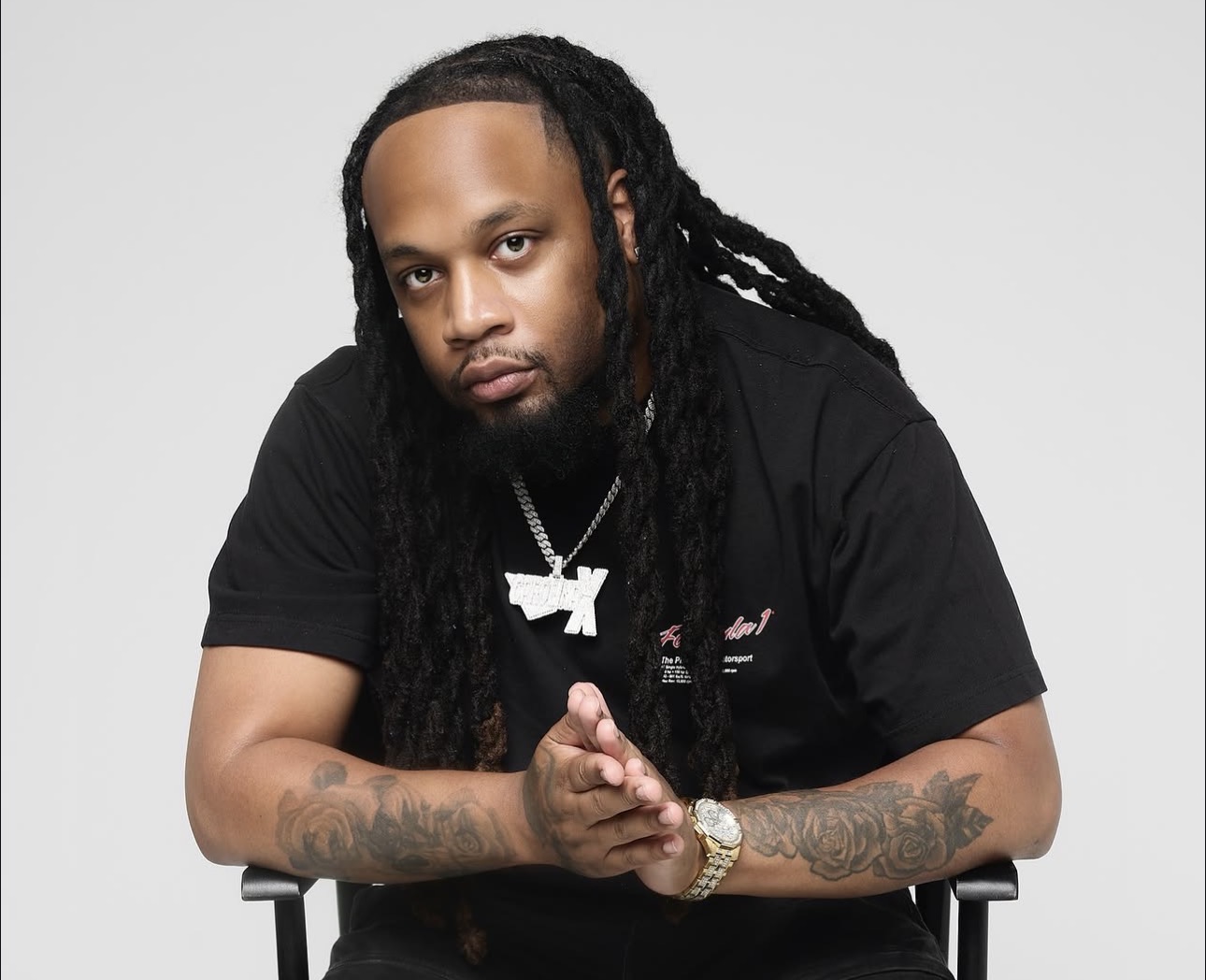 Man with long dreadlocks wearing a black shirt, jewelry, and tattoos, sitting with hands clasped, looking at the camera.