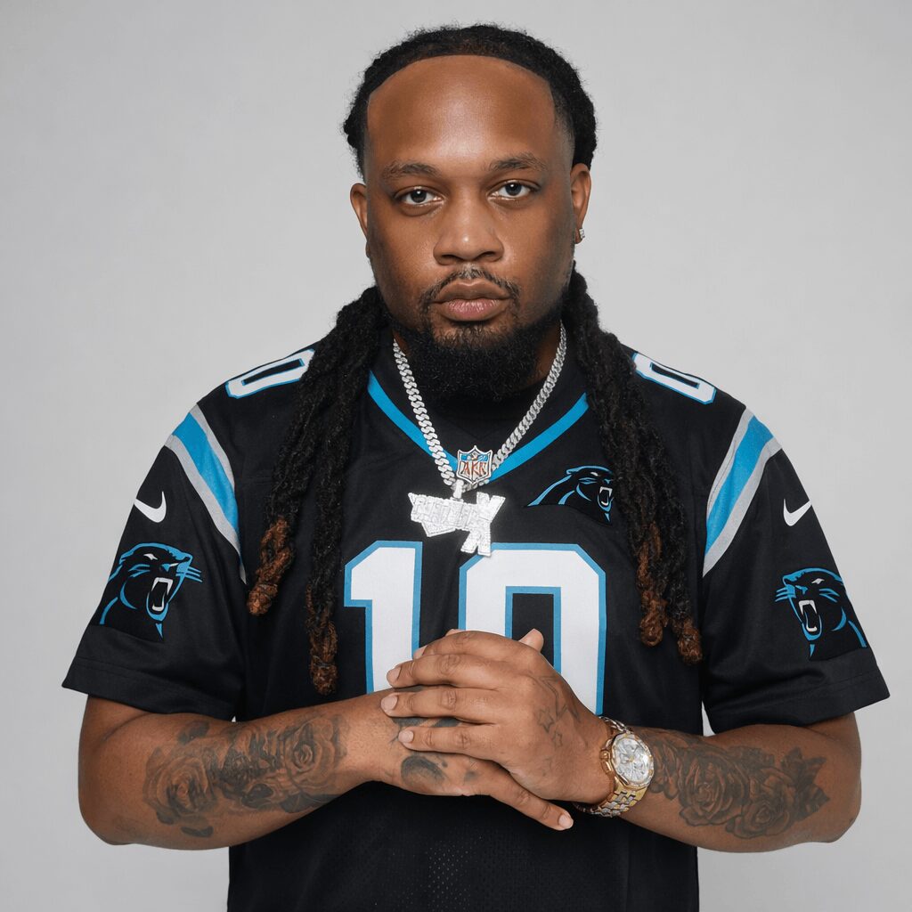 Man with long dreadlocks wearing a black sports jersey with blue and white accents, holding his hands together, against a plain background.