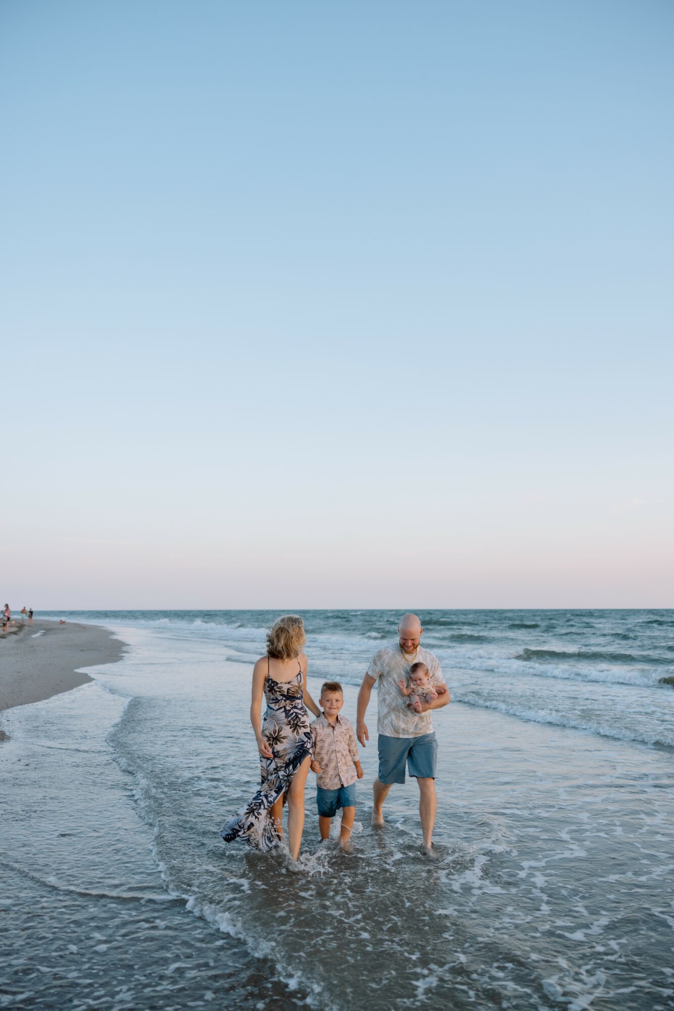 Family of four walking along the shoreline with waves, sky clear, and horizon visible.