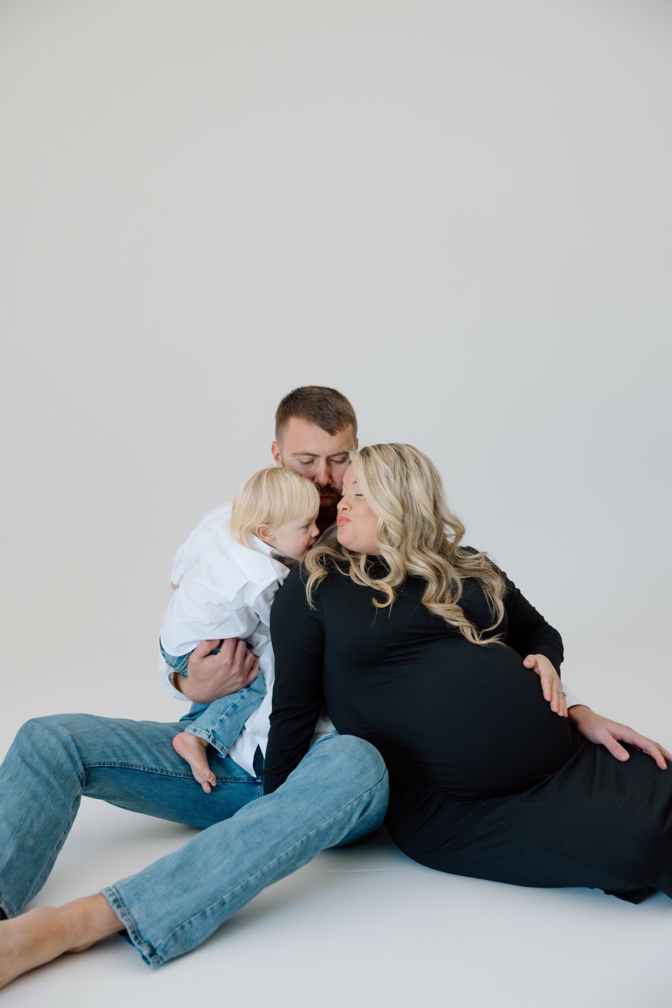 A pregnant woman with long blonde hair lying on the floor, with a man and a child close to her, against a plain light background.