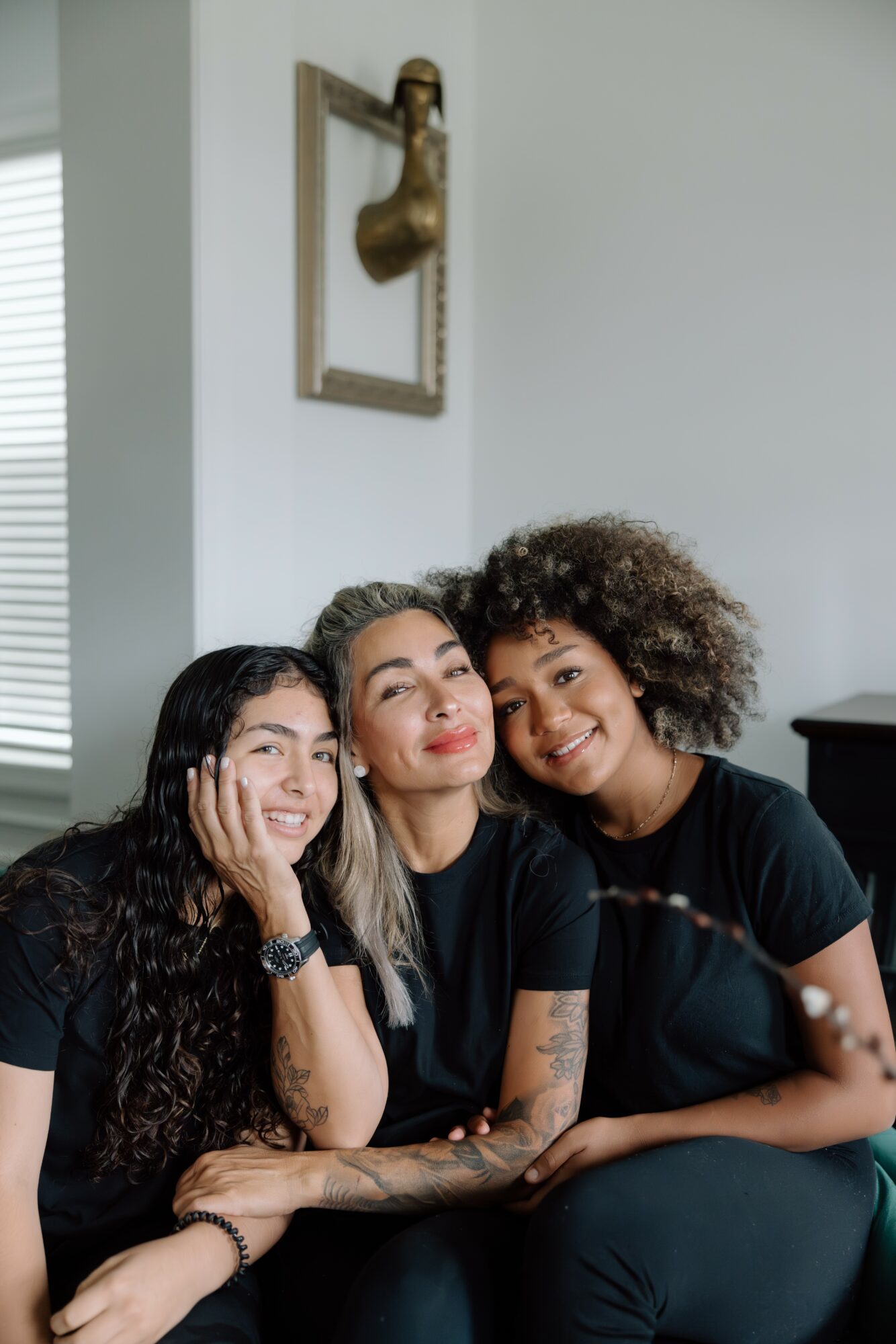 Three women sitting close together indoors, smiling, with a framed decorative object on the wall behind them.