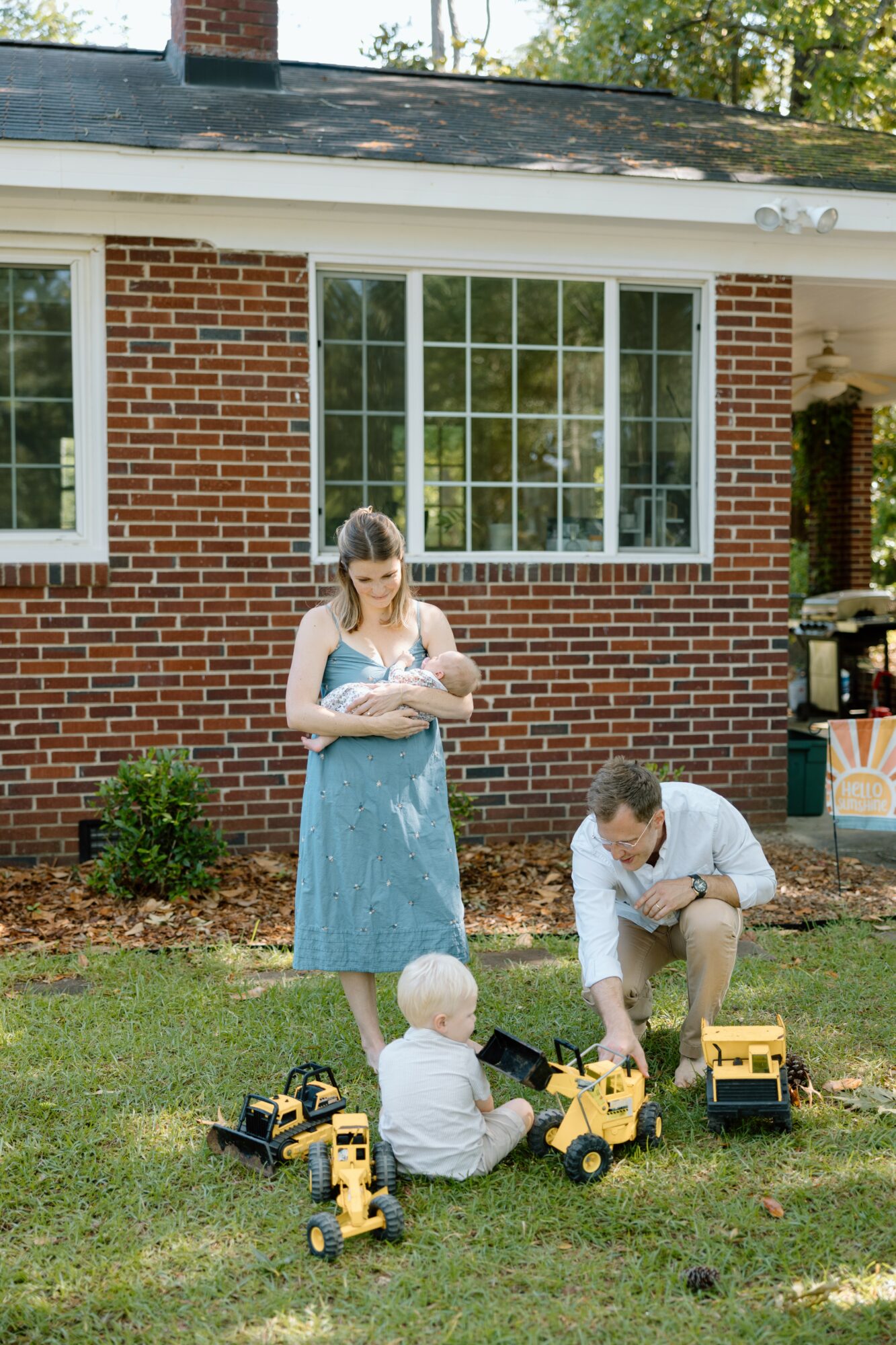 A woman and two children playing with toy construction vehicles on grass in front of a brick house with large windows.