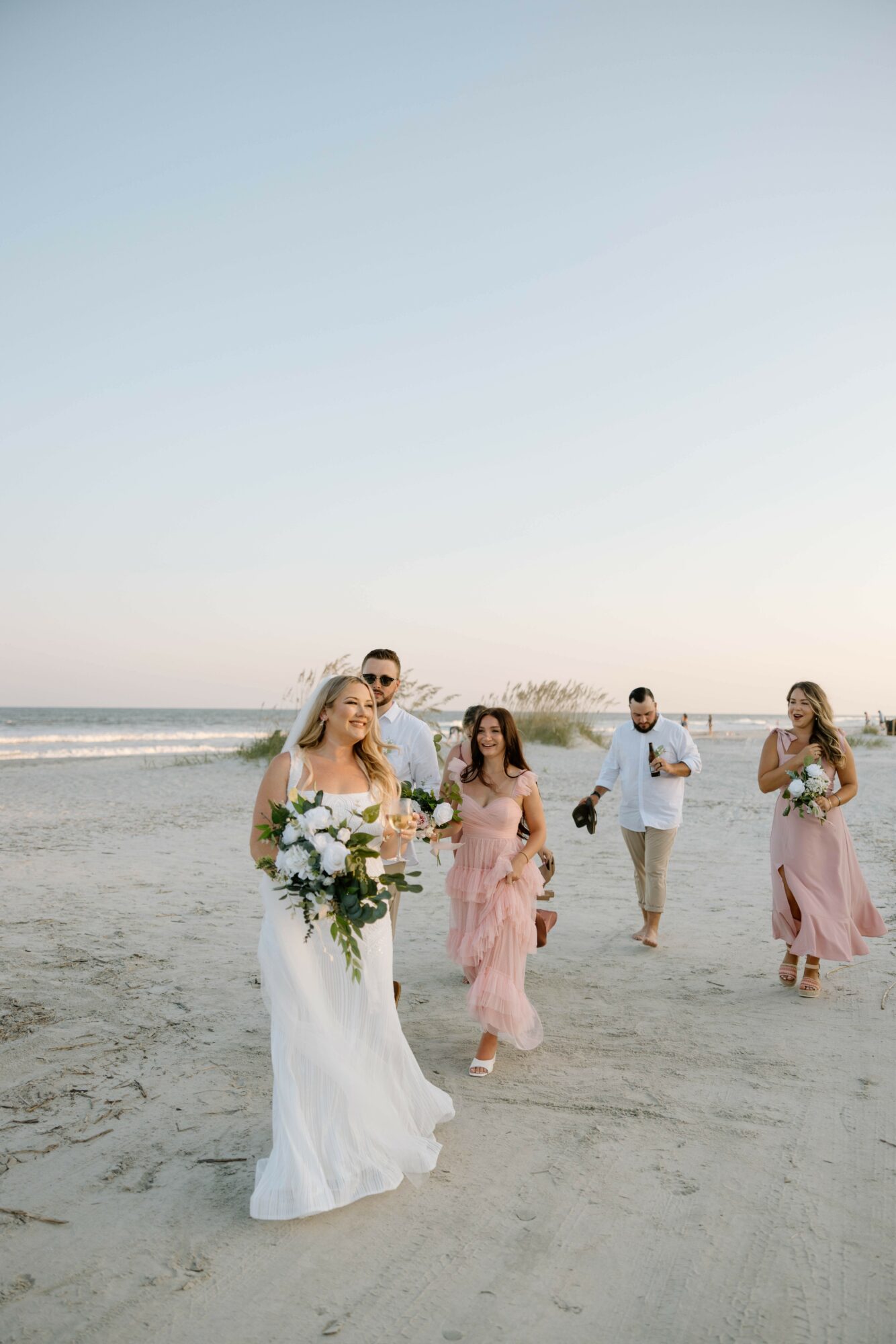 Group of people walking on beach, woman in white dress holding flowers, others in pastel dresses, ocean in background