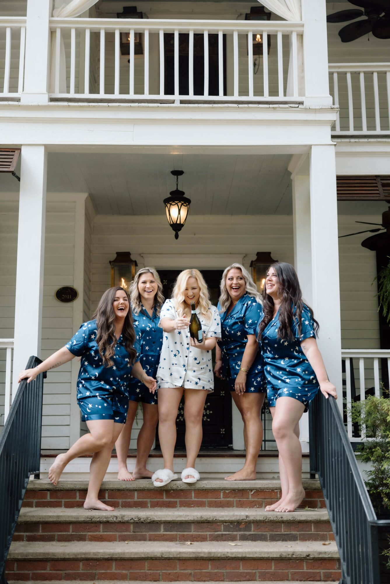 Five women in pajamas standing on stairs outside a house, smiling and posing for a photo, with a porch and hanging light above.