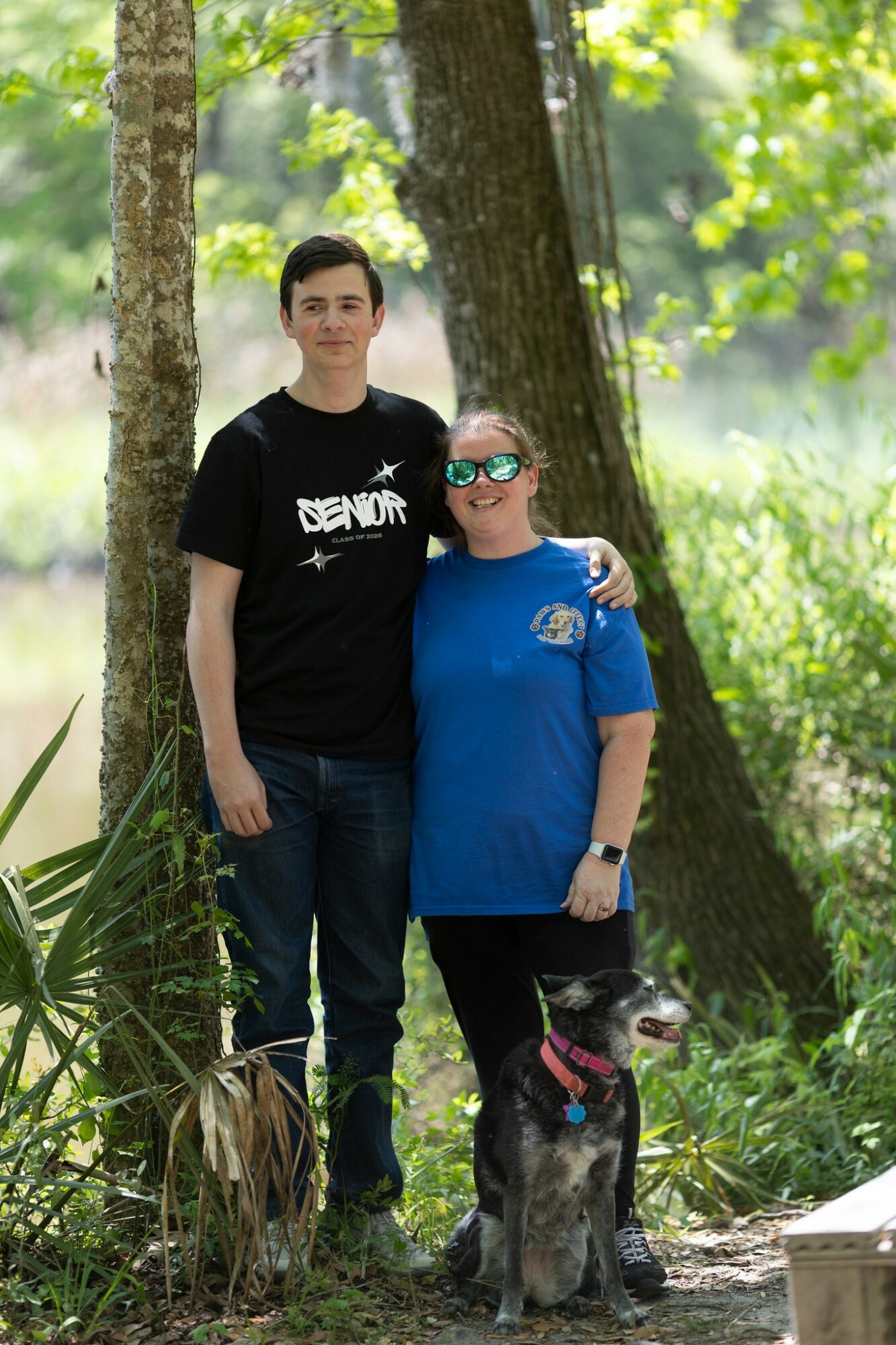 Two people standing outdoors in a forested area, with a dog sitting in front of them, trees and greenery in background.