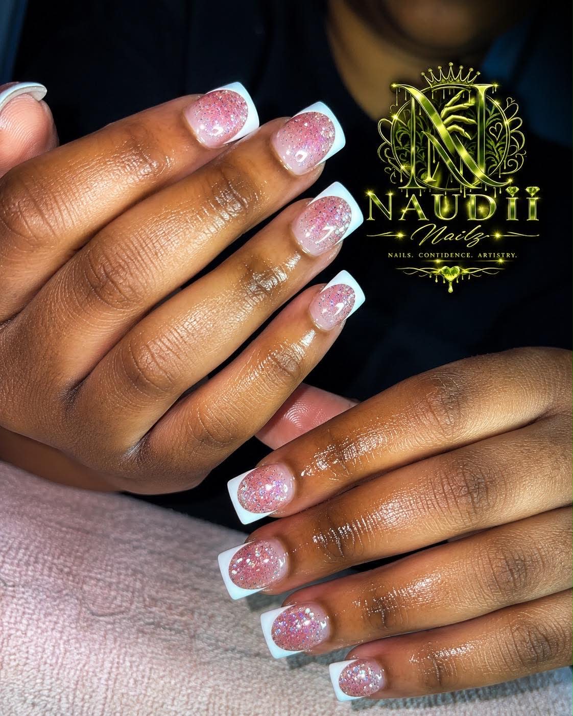 Close-up of hands with decorated nails featuring pink and white glittery designs, resting on a surface.