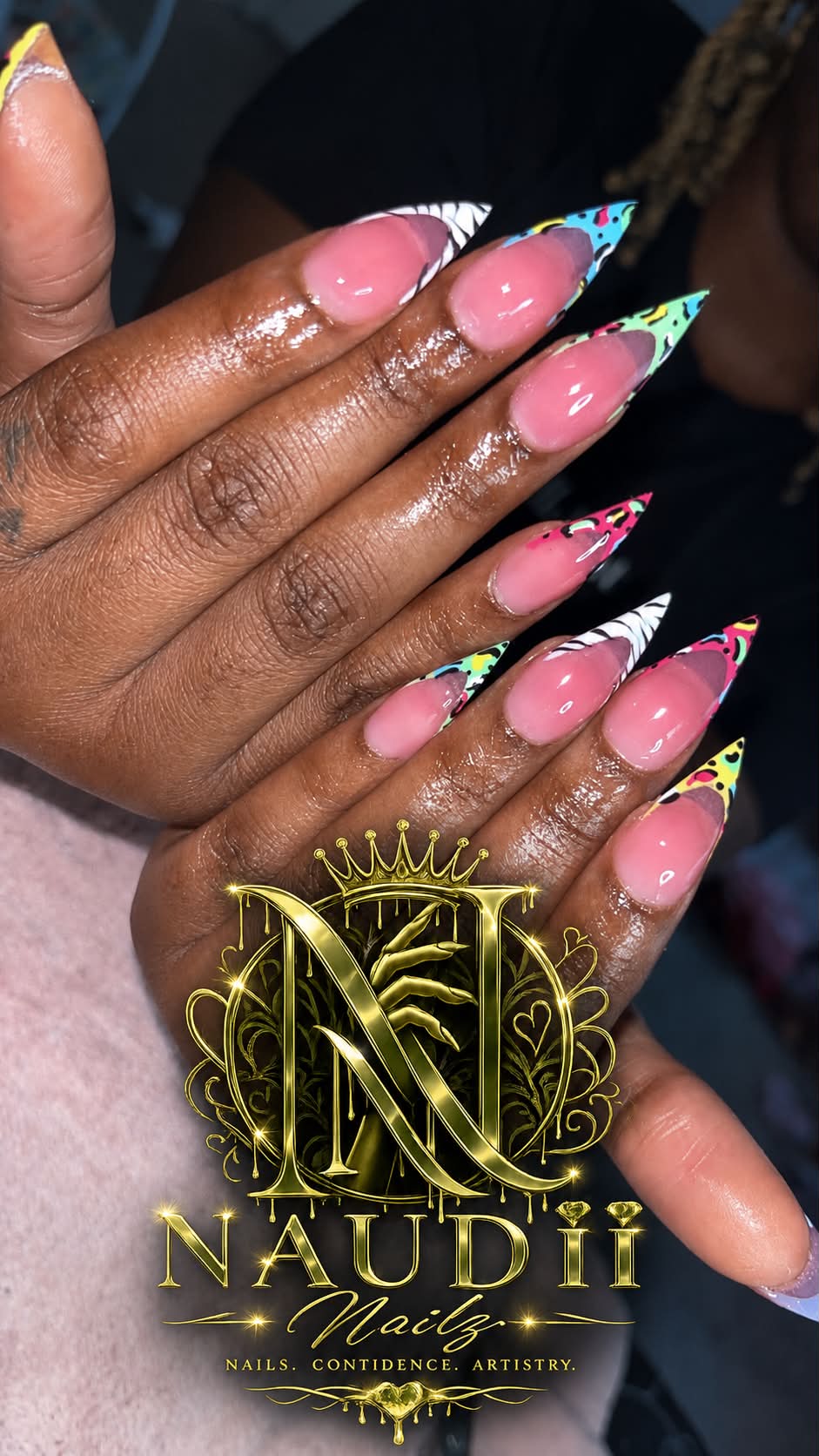 Close-up of hands with long, decorated nails featuring pink, white, and colorful designs, and gold jewelry, against a dark background.