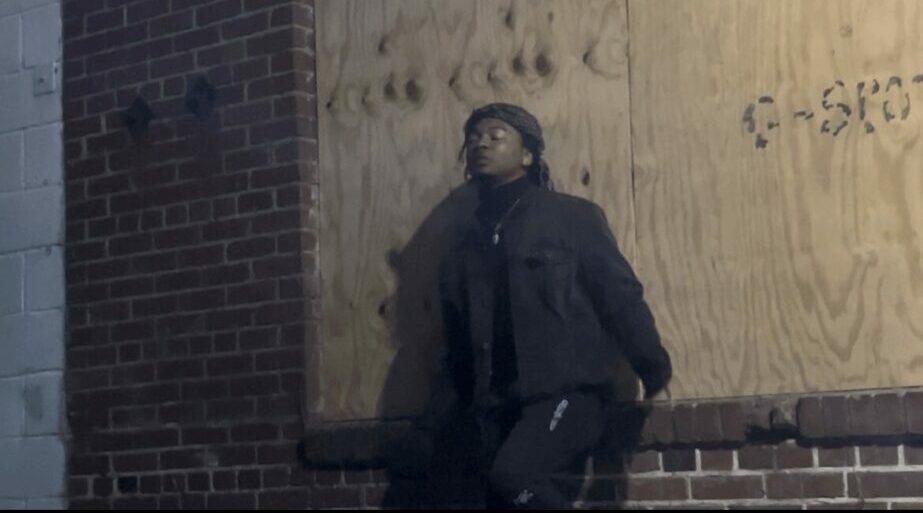 Person wearing a mask and dark clothing leaning against a brick wall in an urban setting.