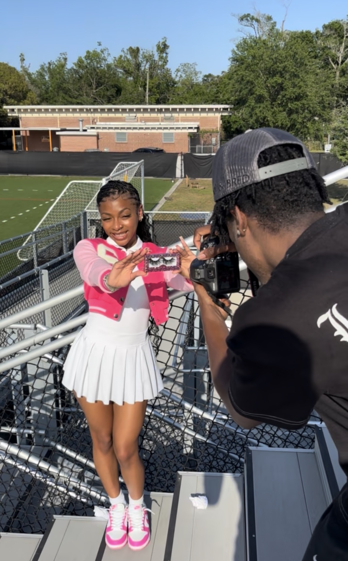 Young girl in pink and white outfit taking a photo with a camera, outdoors on a sports field, with trees in background.