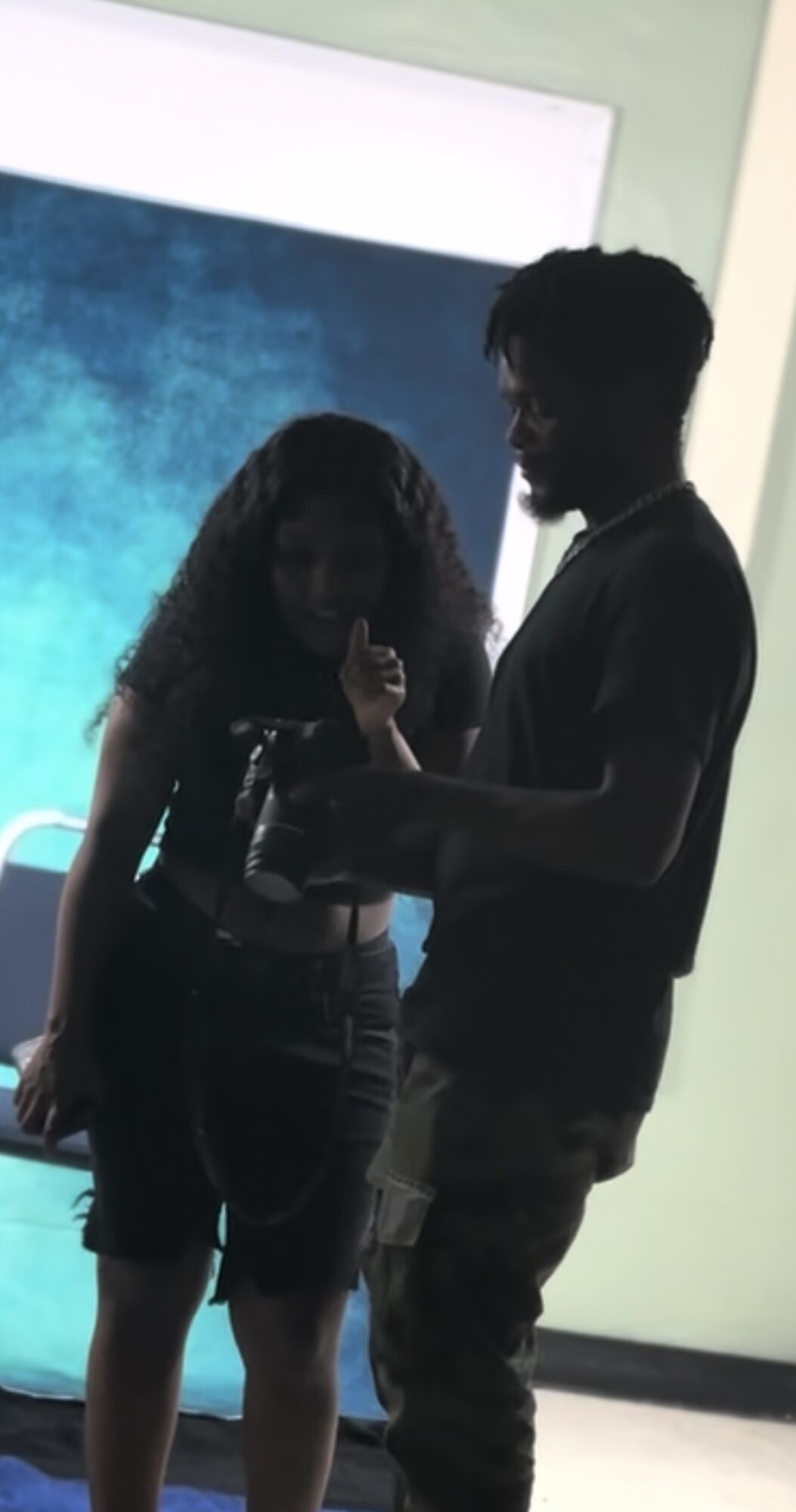 Two people standing indoors, one holding a microphone, with a large screen in the background.