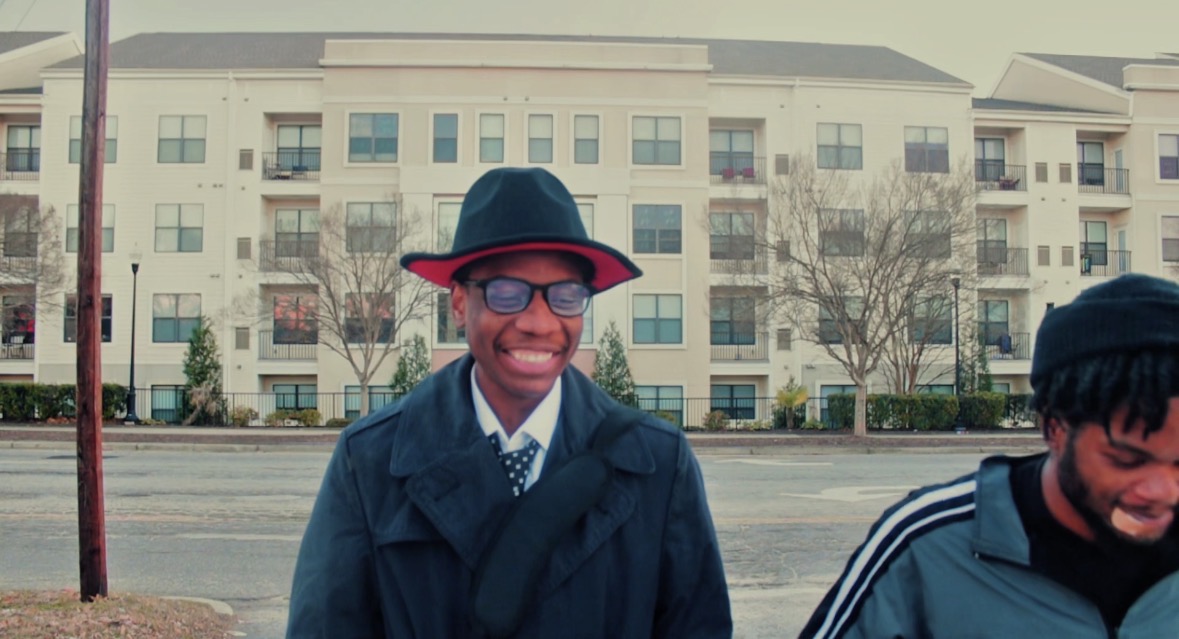 Smiling person wearing glasses, a hat, and a coat, standing outdoors with buildings and trees in background.