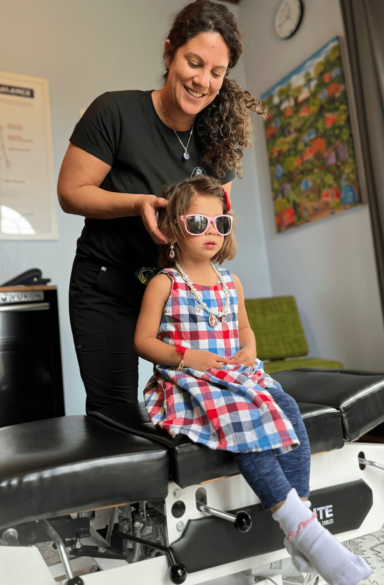 Woman helping girl try on sunglasses in a waiting room, girl sitting on a black bench, colorful artwork on wall behind.