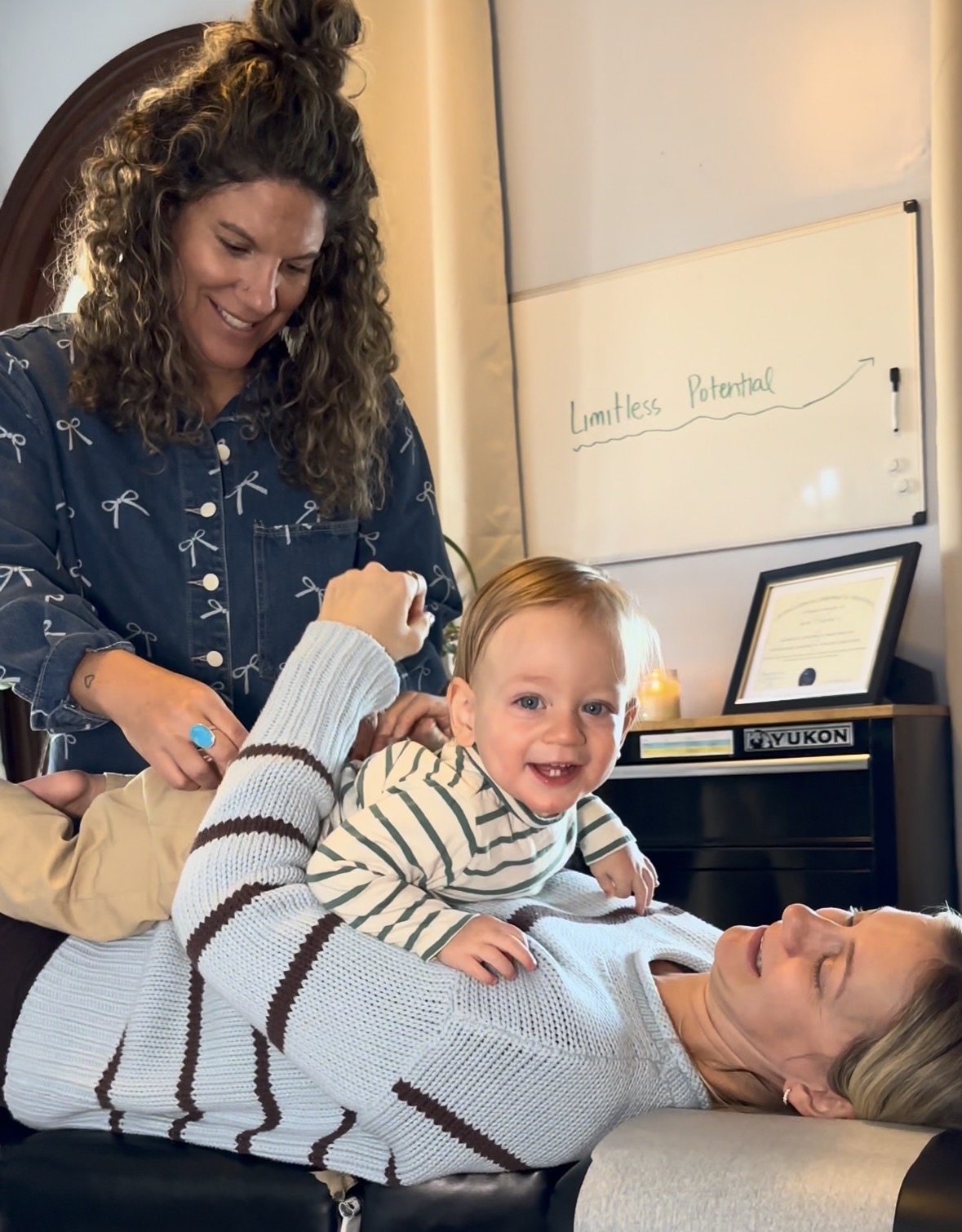 Woman lying on a bed with a smiling child on her stomach, woman standing nearby smiling, in a room with a whiteboard and framed certificate.