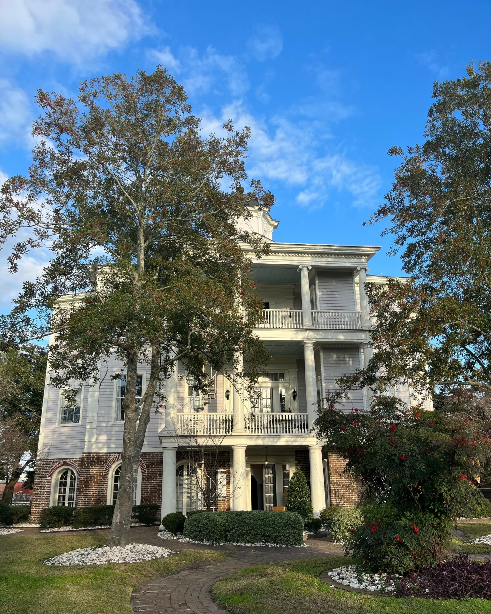 Three-story house with balconies, surrounded by trees and a landscaped yard, under a blue sky with clouds.