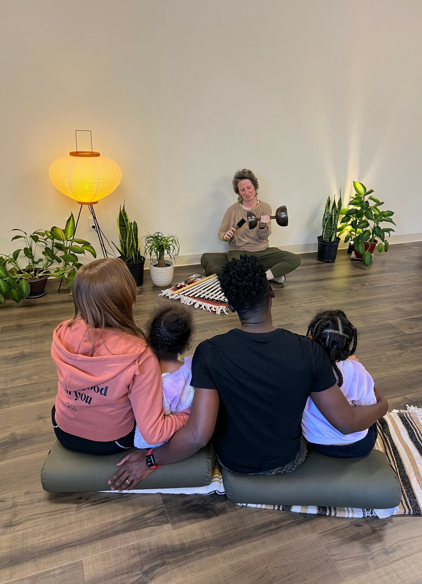 Person sitting on the floor with a microphone, speaking to a group of children and an adult, in a room with plants and a lamp.