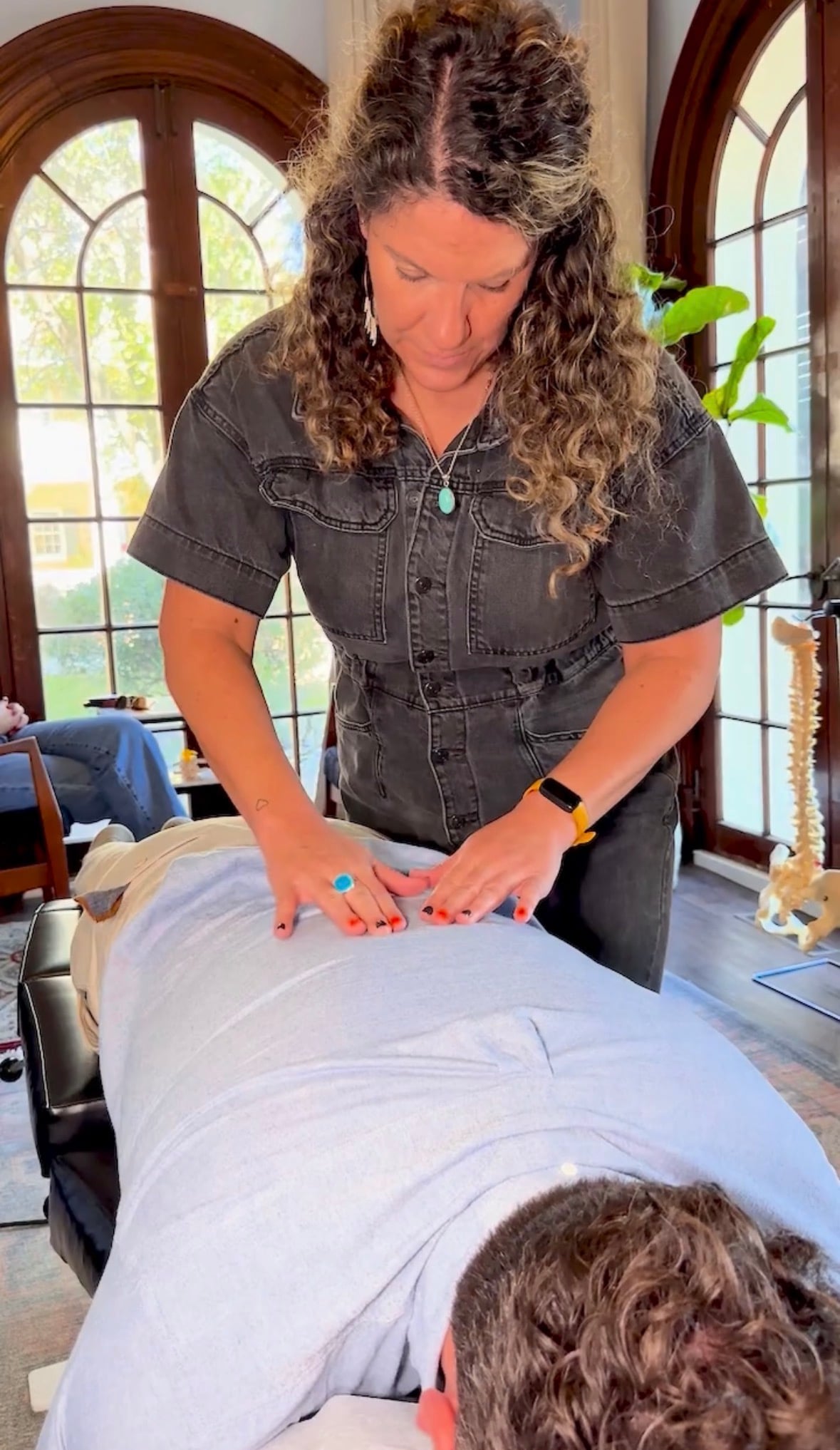 Woman giving a massage or therapy to a person lying face down on a bed or table, in a room with large windows and plants.