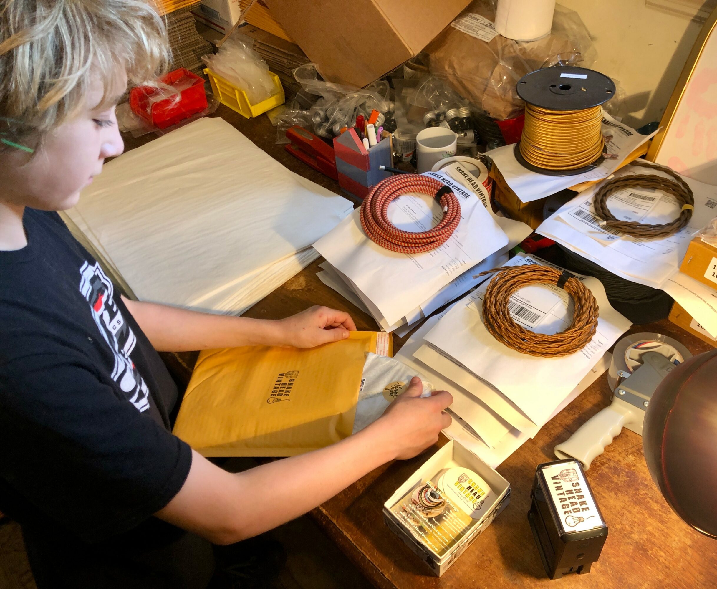 Child working at a cluttered desk with various craft supplies, including spools of wire and a yellow package.