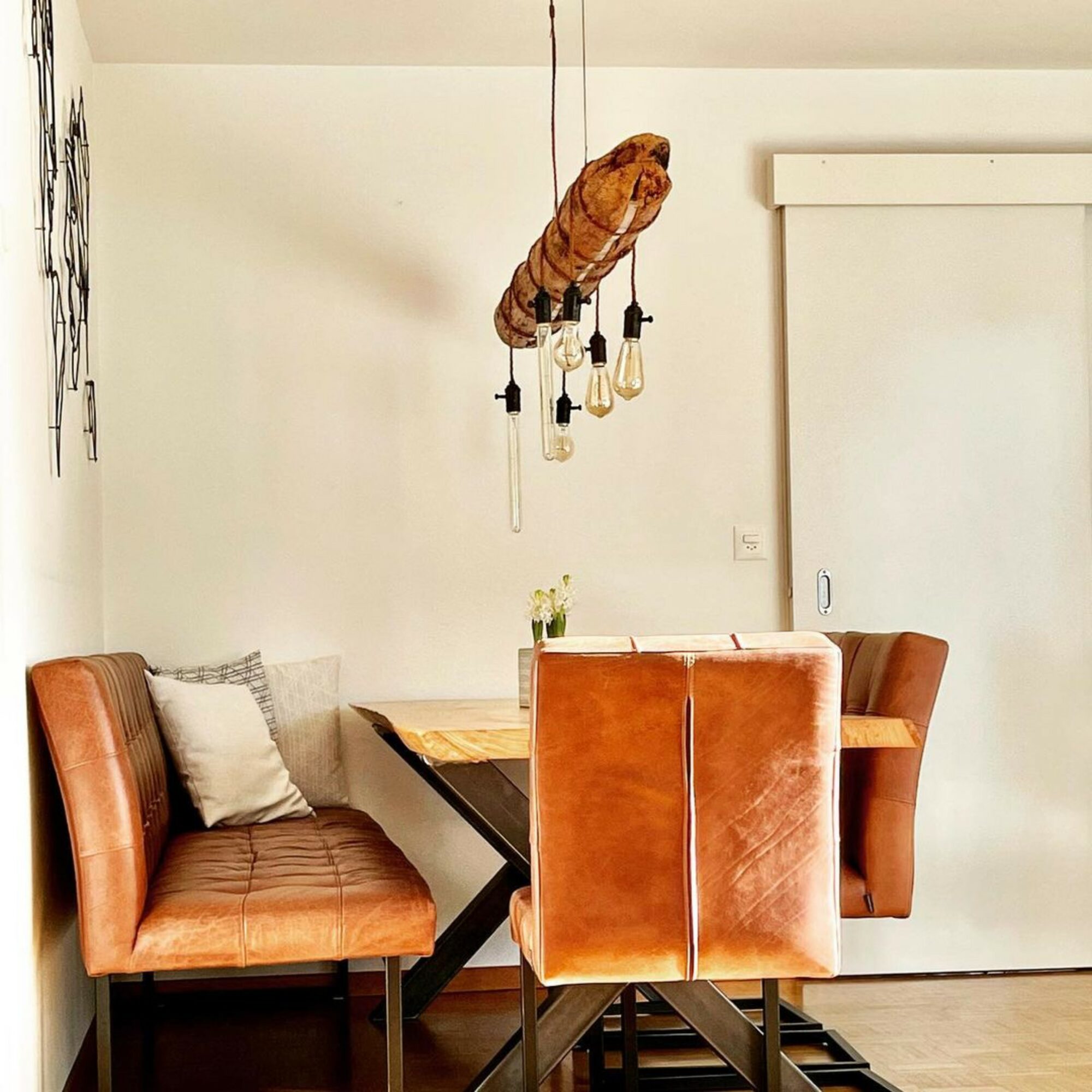 Dining area with a wooden table, four leather chairs, and a hanging light fixture with exposed bulbs, against a white wall.