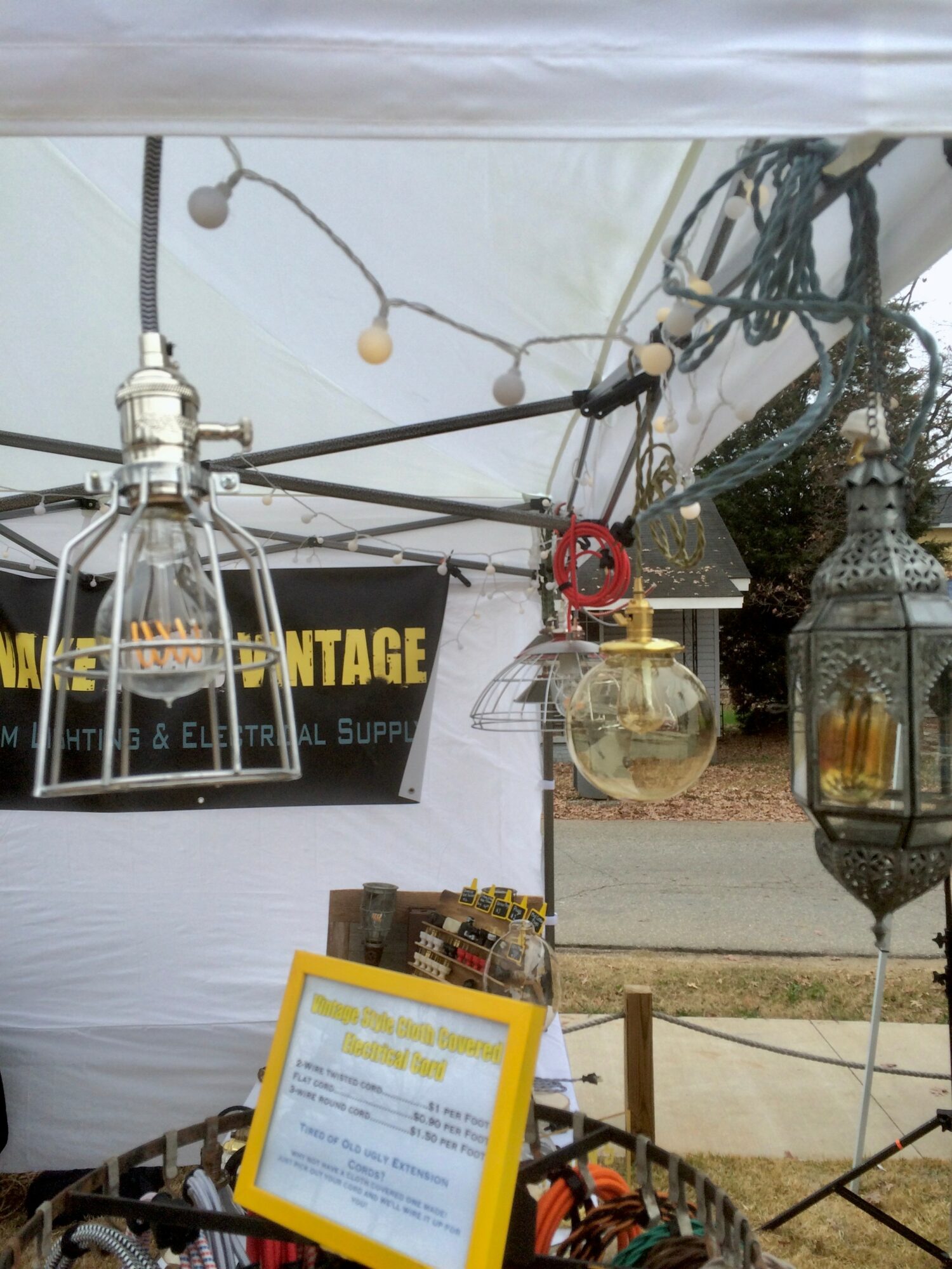 Outdoor scene with hanging lanterns, string lights, and a sign, under a white canopy. A basket with items is in the foreground.