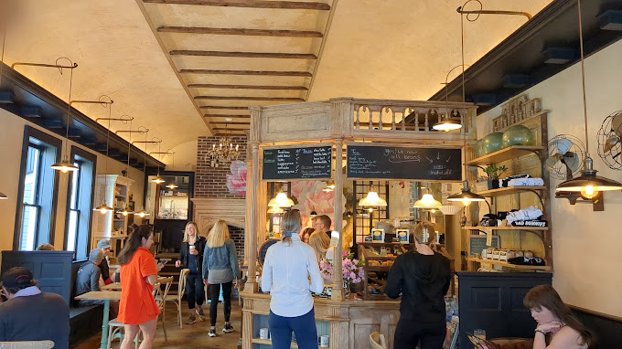 People inside a cafe or restaurant, some standing and some sitting, with a counter and shelves behind it, warm lighting.