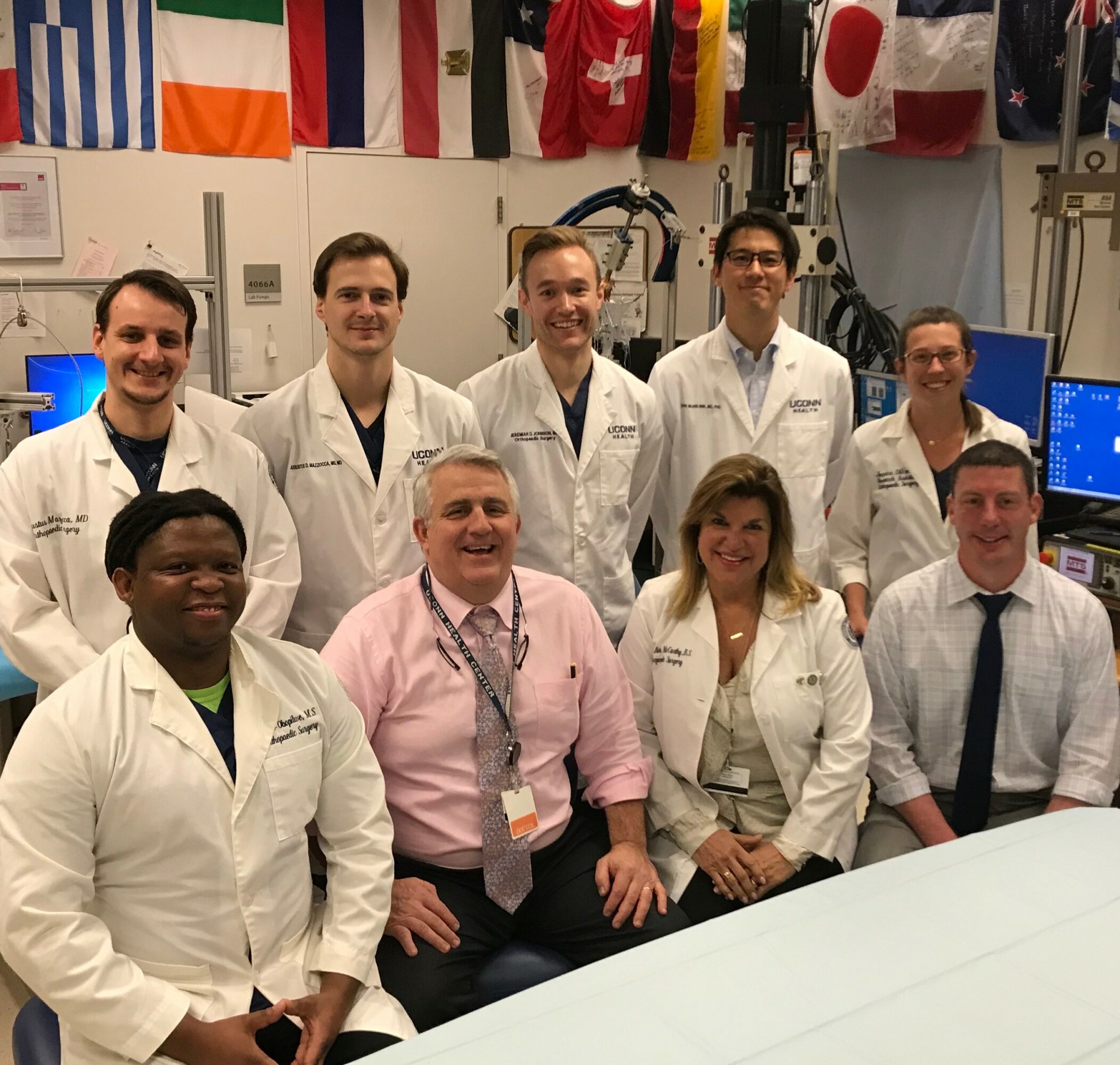 Group of nine people in lab coats in a room with international flags and medical equipment, smiling at the camera.