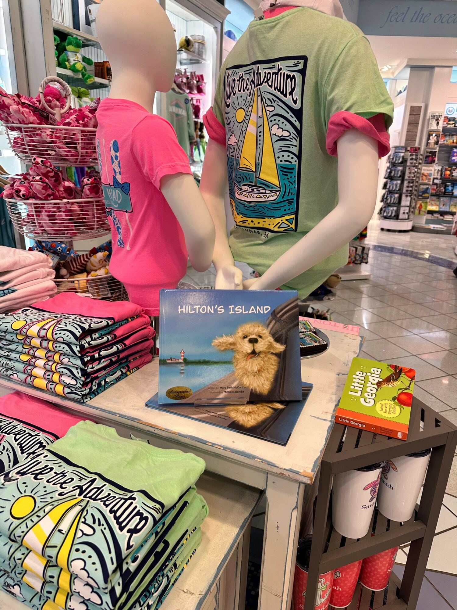 Two children with light skin and long hair look at a small puppy in a box on a table in a store. One child wears a green shirt, the other a pink shirt. The store has shelves with various products. The puppy is light brown and fluffy.