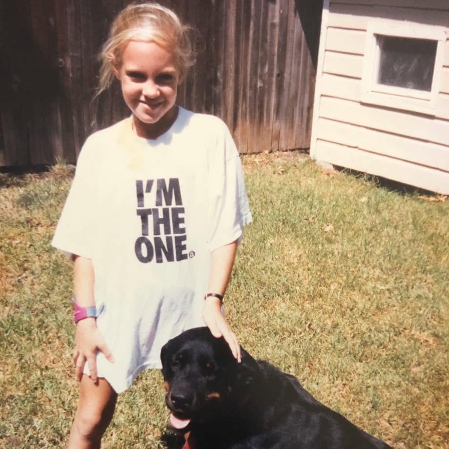 Young girl with blonde hair smiling outdoors, standing next to a black dog on grass, with a wooden fence and house in background.