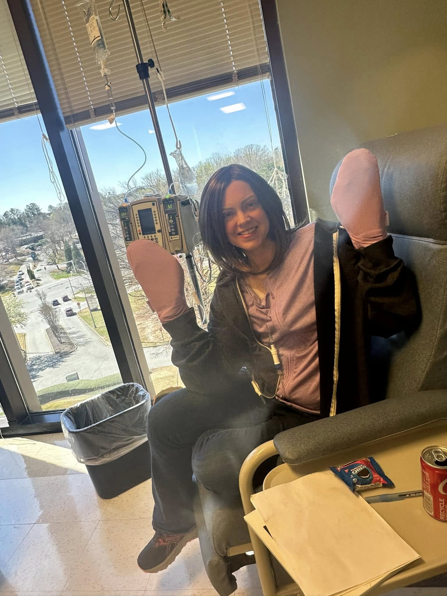 Person sitting in a hospital room with IV drip, smiling, window showing outdoor scene, medical equipment nearby.
