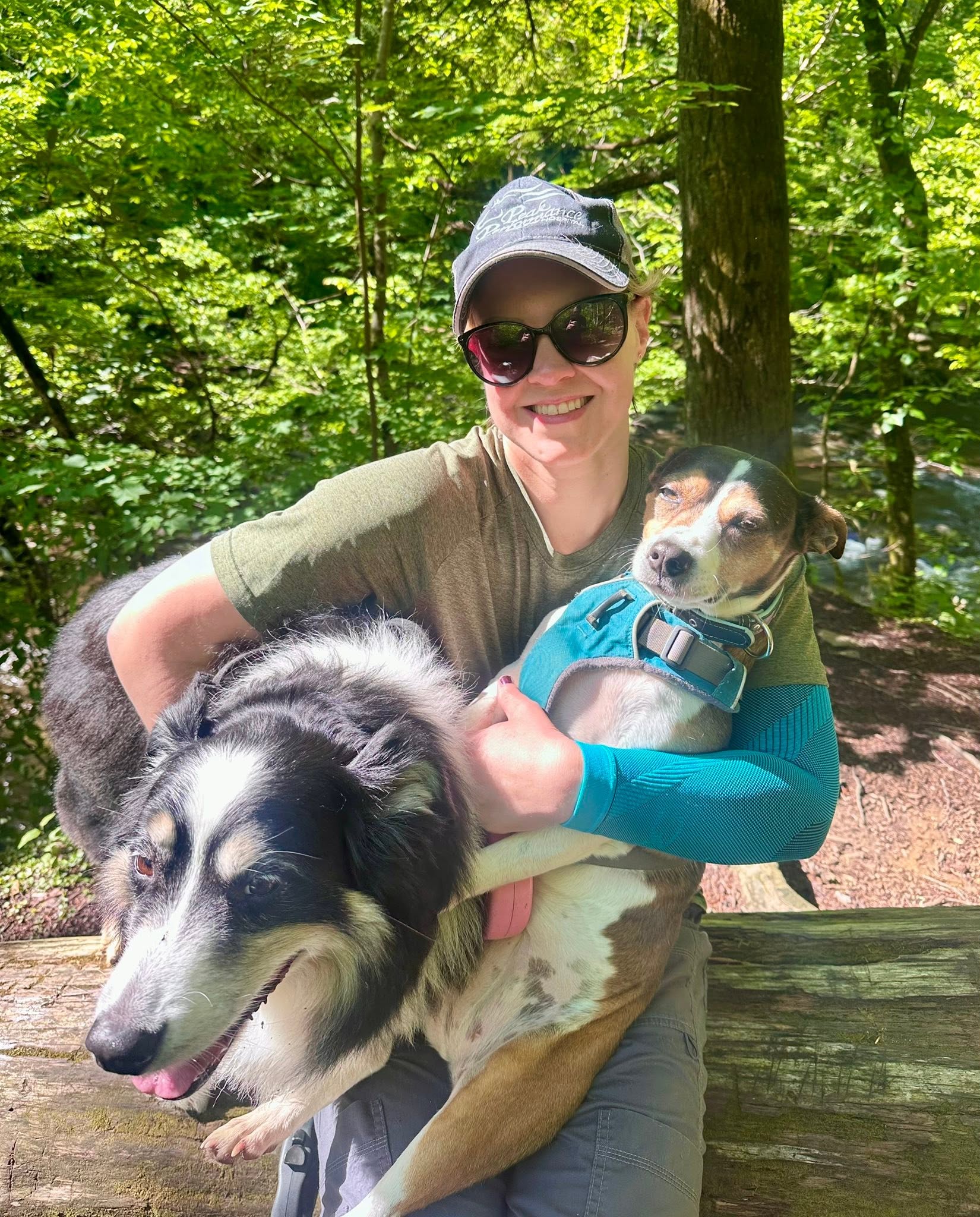 Woman in sunglasses and cap holding two dogs outdoors in a forested area, smiling.