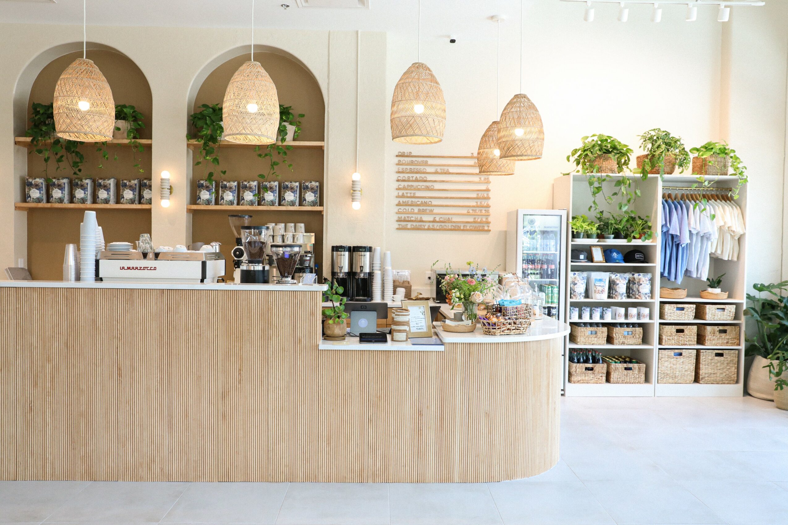 Coffee shop counter with hanging pendant lights, shelves with plants and products, and a display fridge, in a bright space.