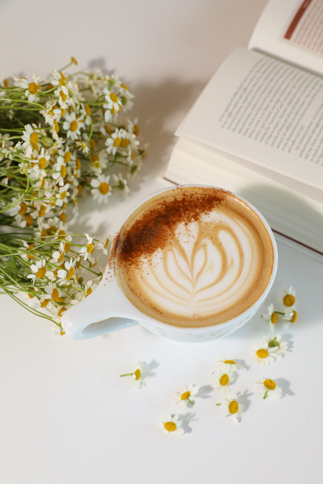 Cup of coffee with latte art, daisies on a white surface, open book nearby, soft lighting