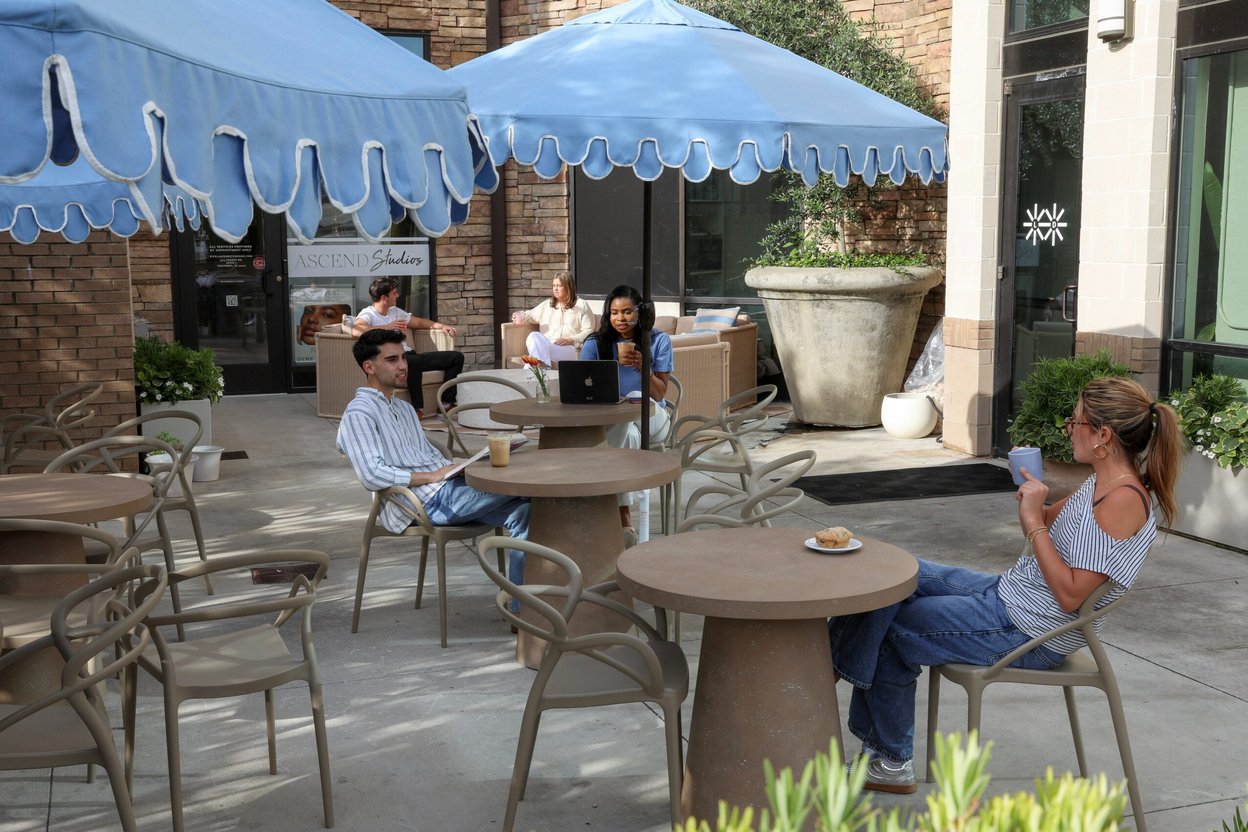 People sitting at outdoor tables under blue umbrellas on a patio, some using phones, with a large planter nearby.