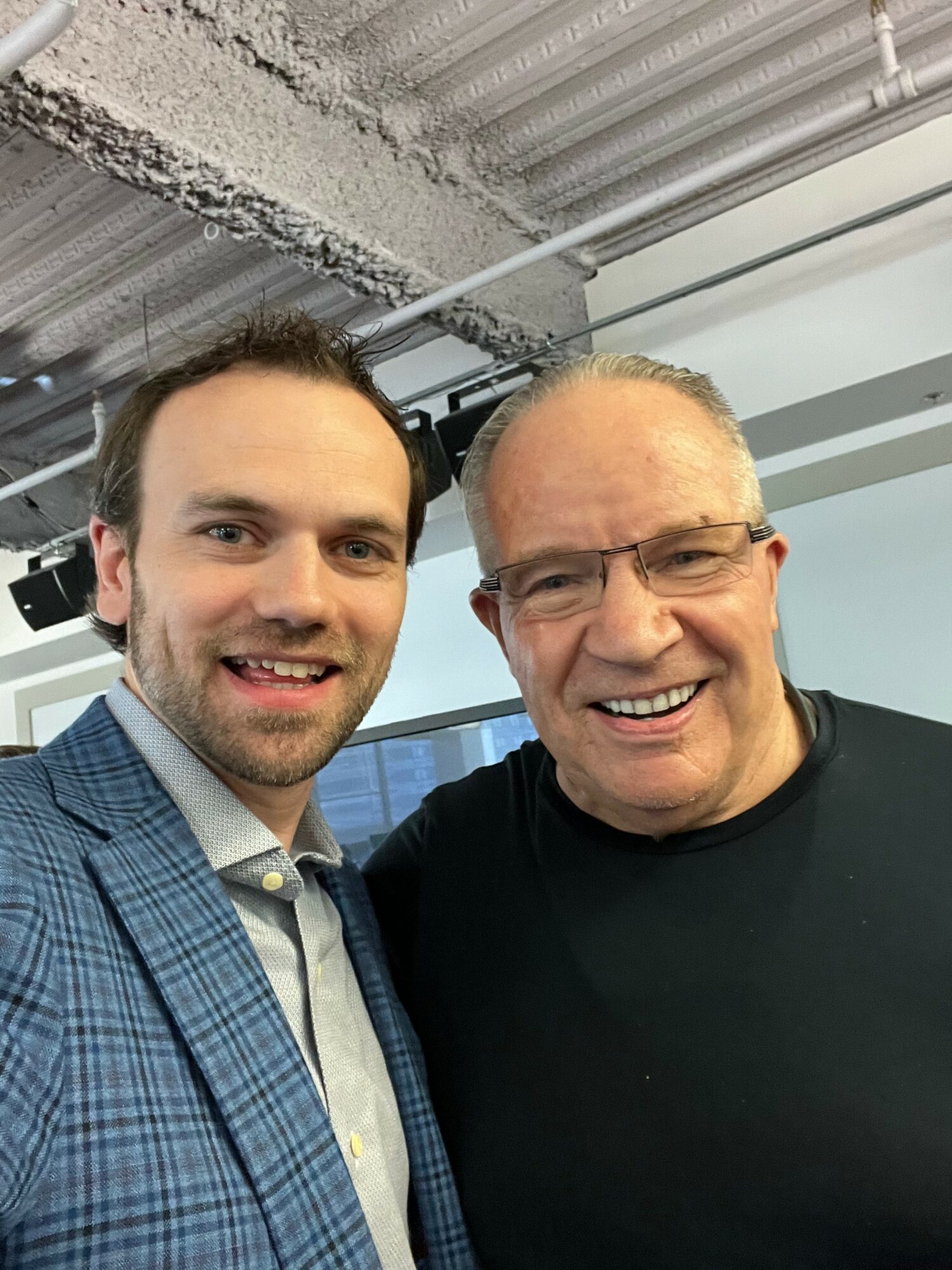 Two men smiling, one wearing a blue checkered suit and the other in a black shirt, indoors with a concrete ceiling.