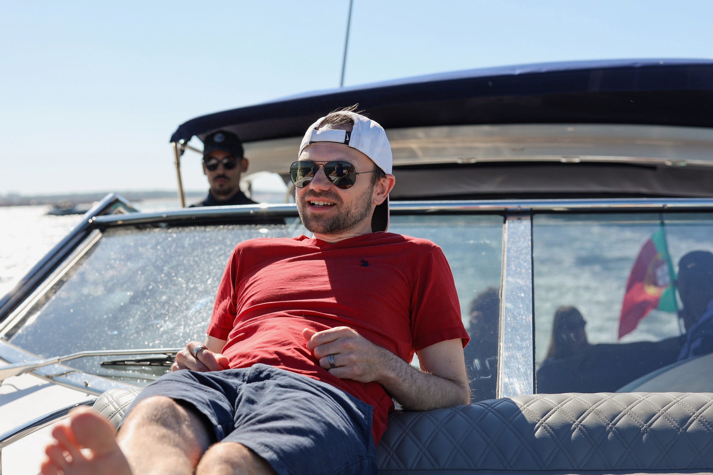 Man in red shirt and white cap relaxing on boat, with another person in background, sunny day.