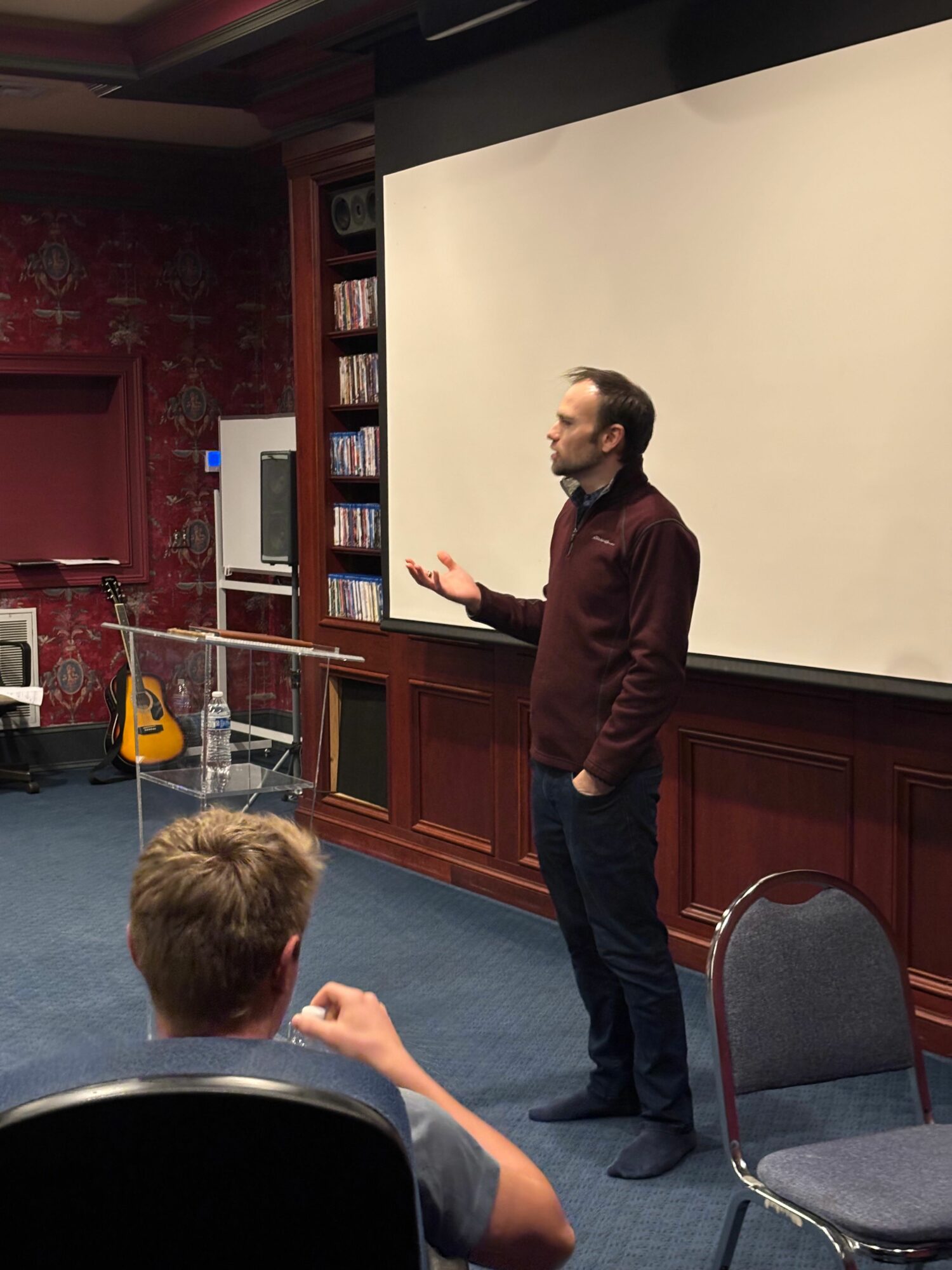 Person standing and gesturing in front of a large whiteboard in a room with bookshelves and chairs.