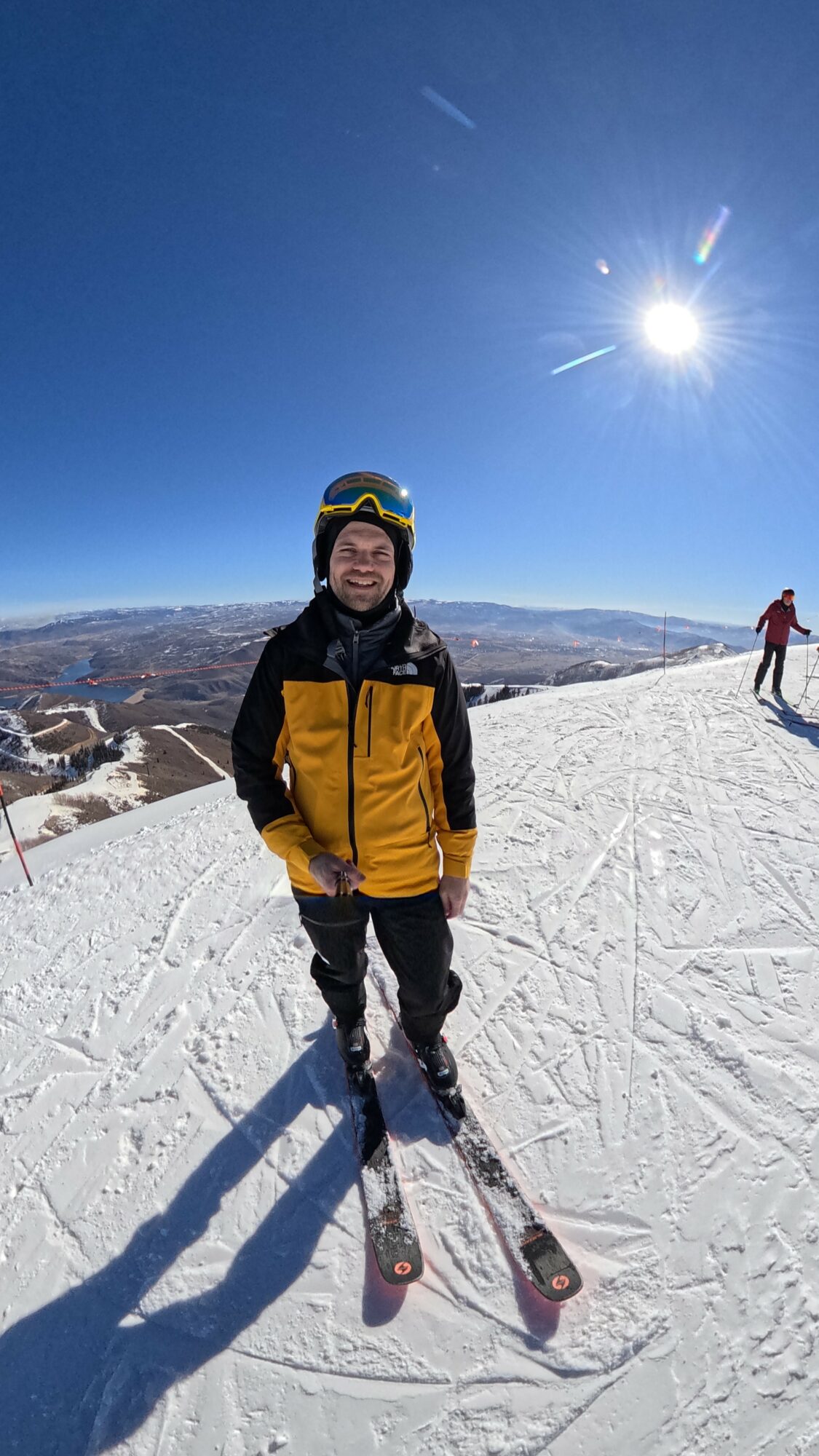 Child in yellow and black ski gear standing on snow with skis, bright sun in clear blue sky, distant mountains, and another person in background.