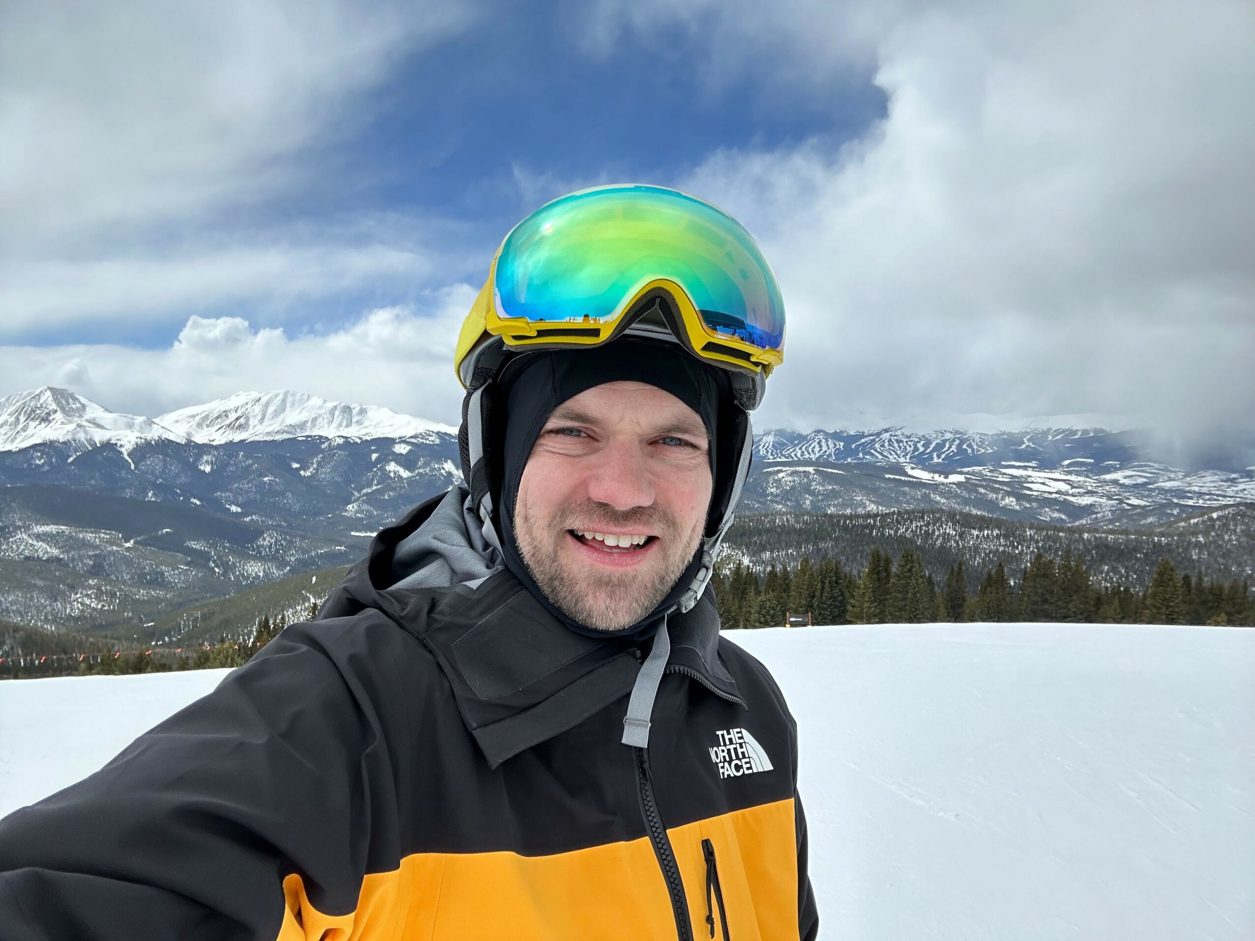 Person wearing ski gear and goggles, smiling, with snowy landscape in background.