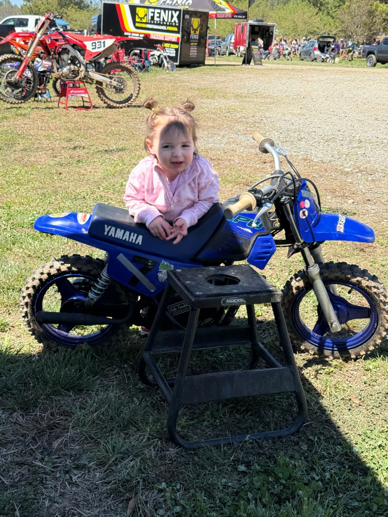 Child sitting on a blue Yamaha mini motorcycle with a black chair in front, outdoors on grass, with bicycles in background.