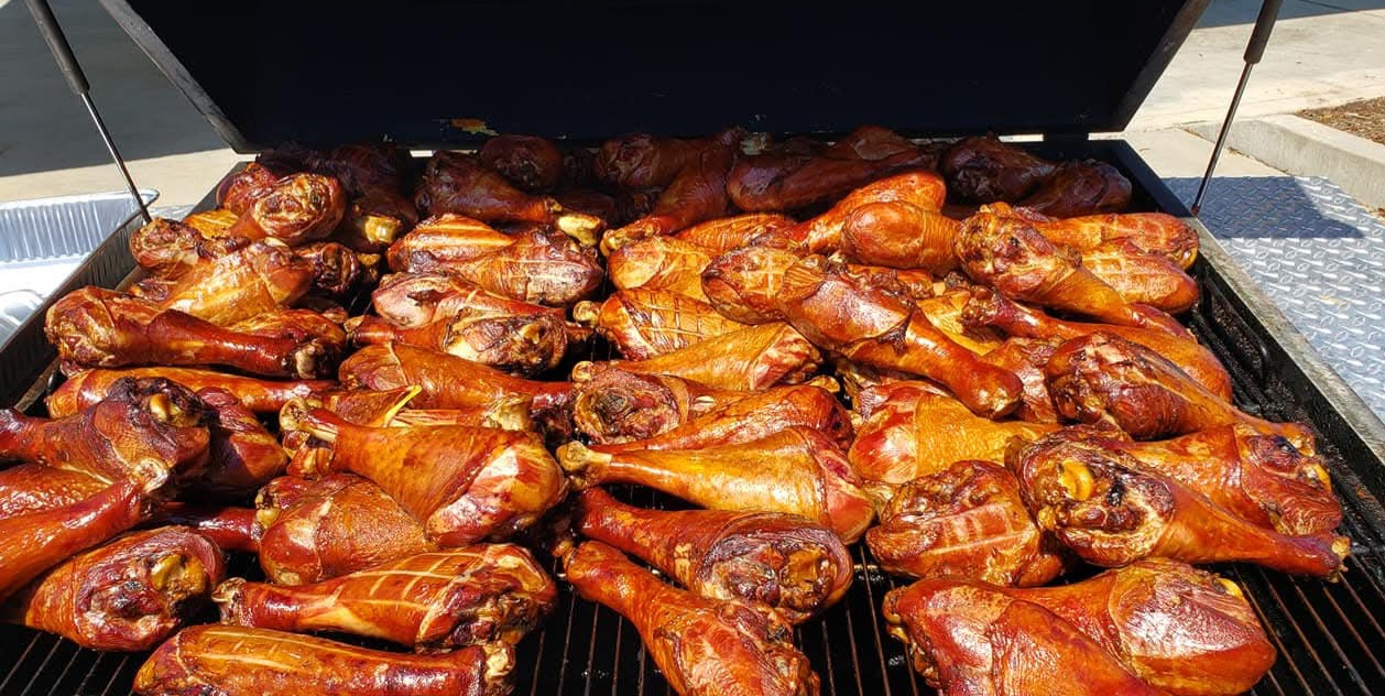 Barbecue grill with numerous cooked chicken drumsticks and pieces, some with charred skin, arranged on the grill surface.