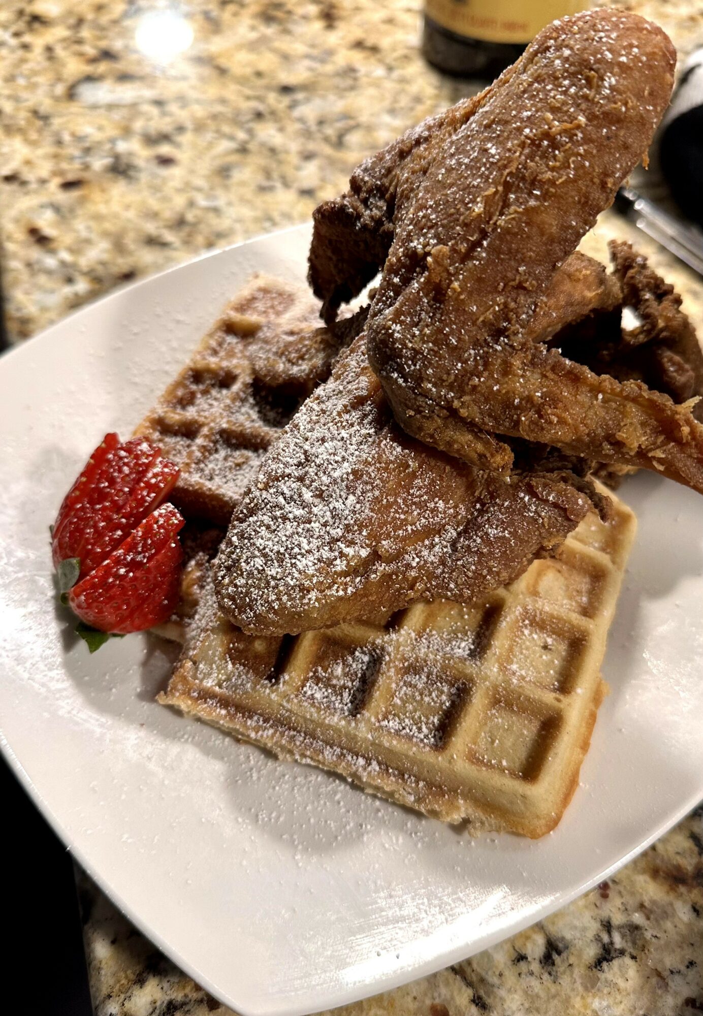 Plate with waffle, fried chicken, and strawberries on a white dish, on a granite countertop.